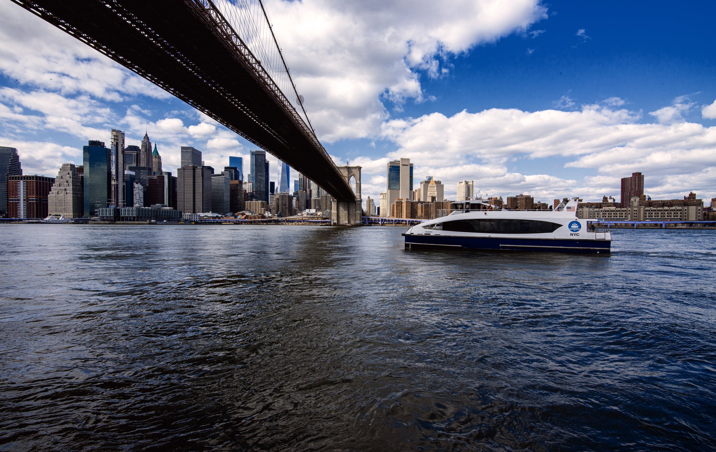 View of New York City skyline and Brooklyn Bridge from the water, with a white and black boat labeled NYC Ferry in the foreground, and the sky filled with clouds.