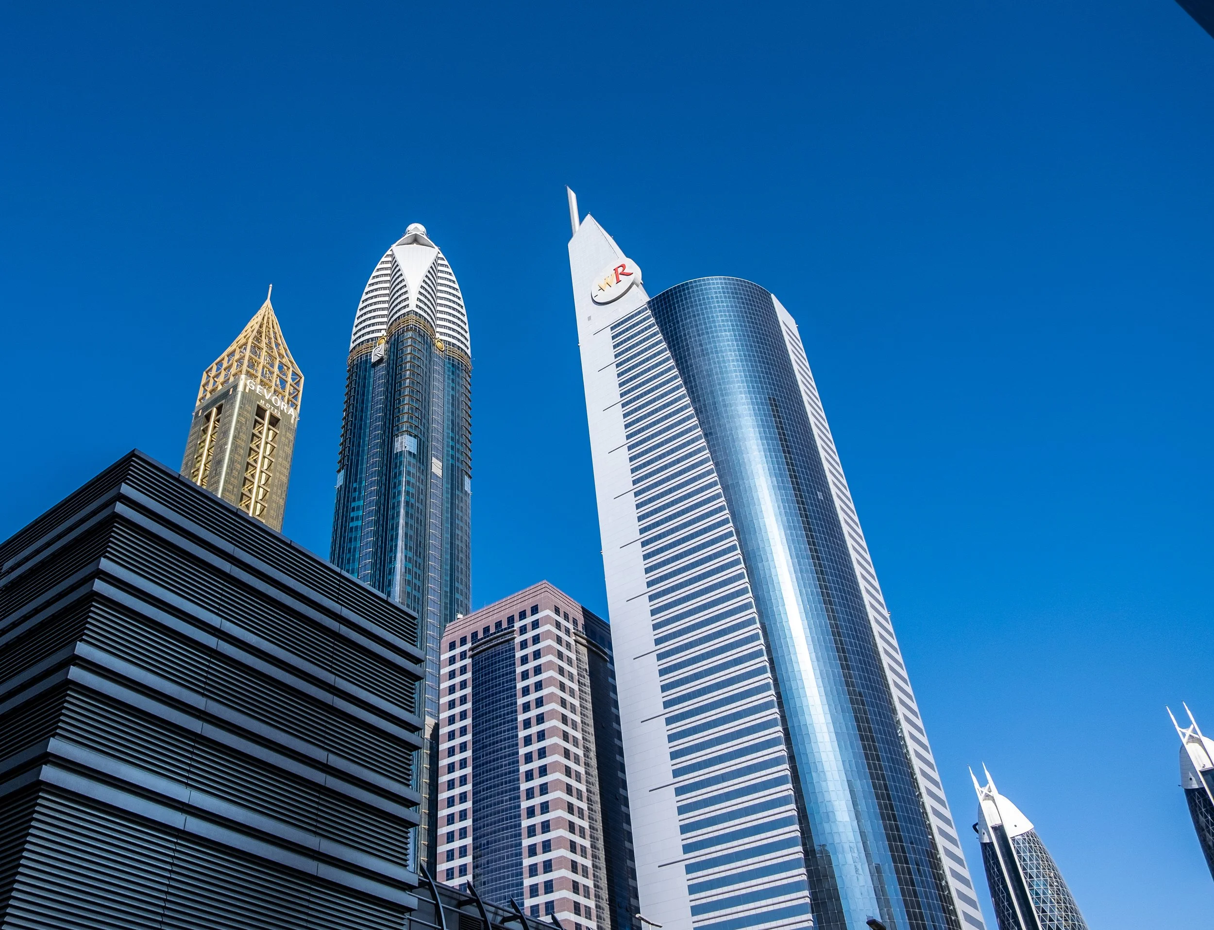 A view of tall modern skyscrapers in a city against a clear blue sky, featuring glass and steel buildings with distinctive shapes and designs.