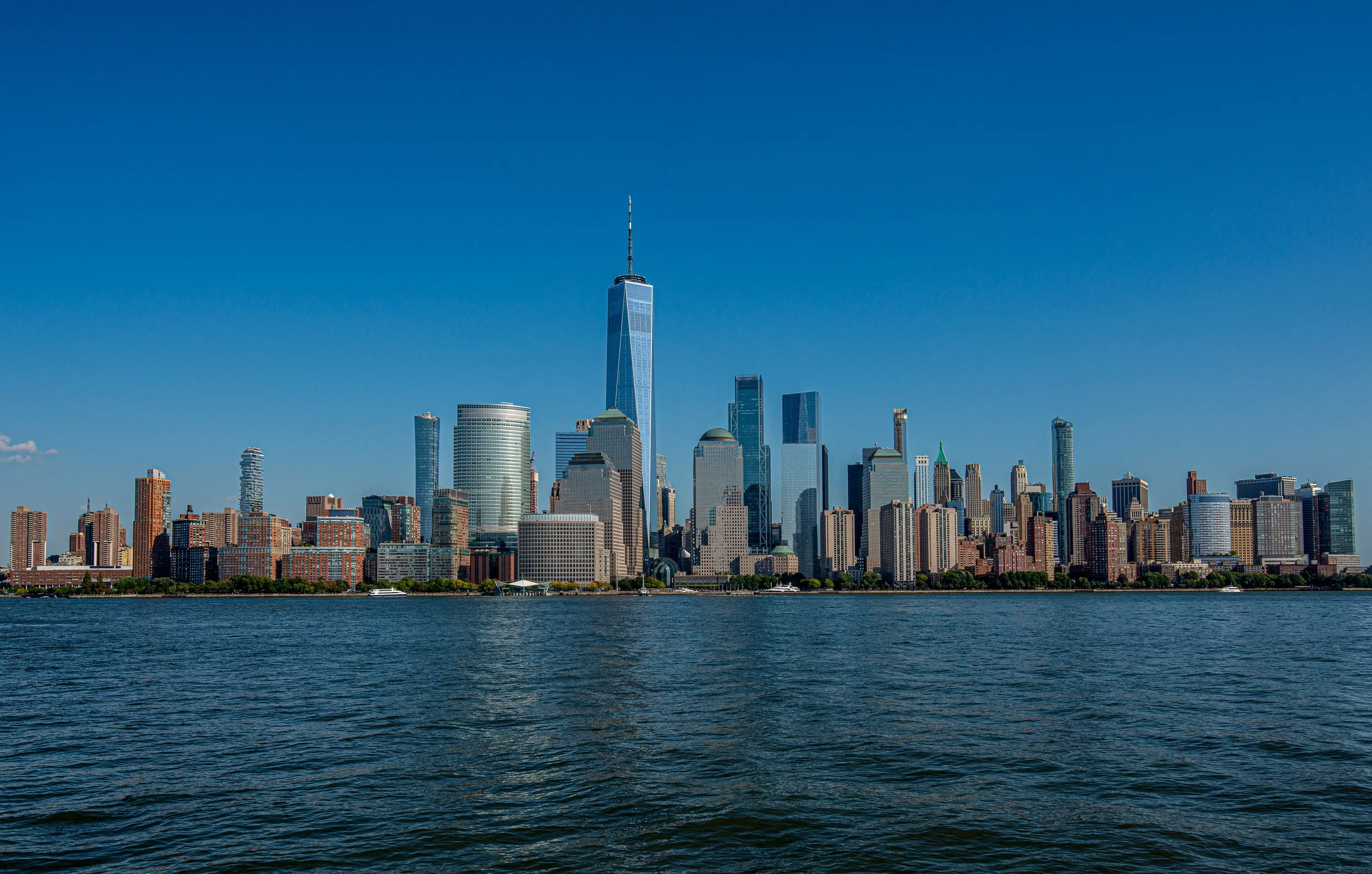 A panoramic view of the New York City skyline from across the water on a clear, sunny day, featuring One World Trade Center and various skyscrapers.