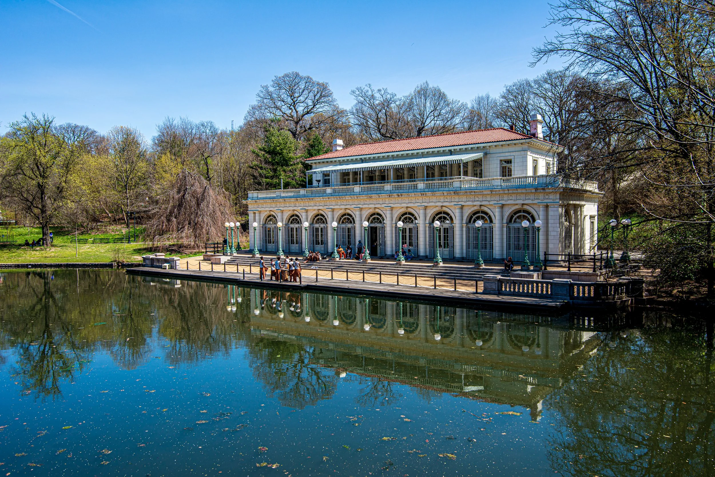 A historic white building with arched windows and balconies, located on the edge of a pond with trees in the background, under a clear blue sky.