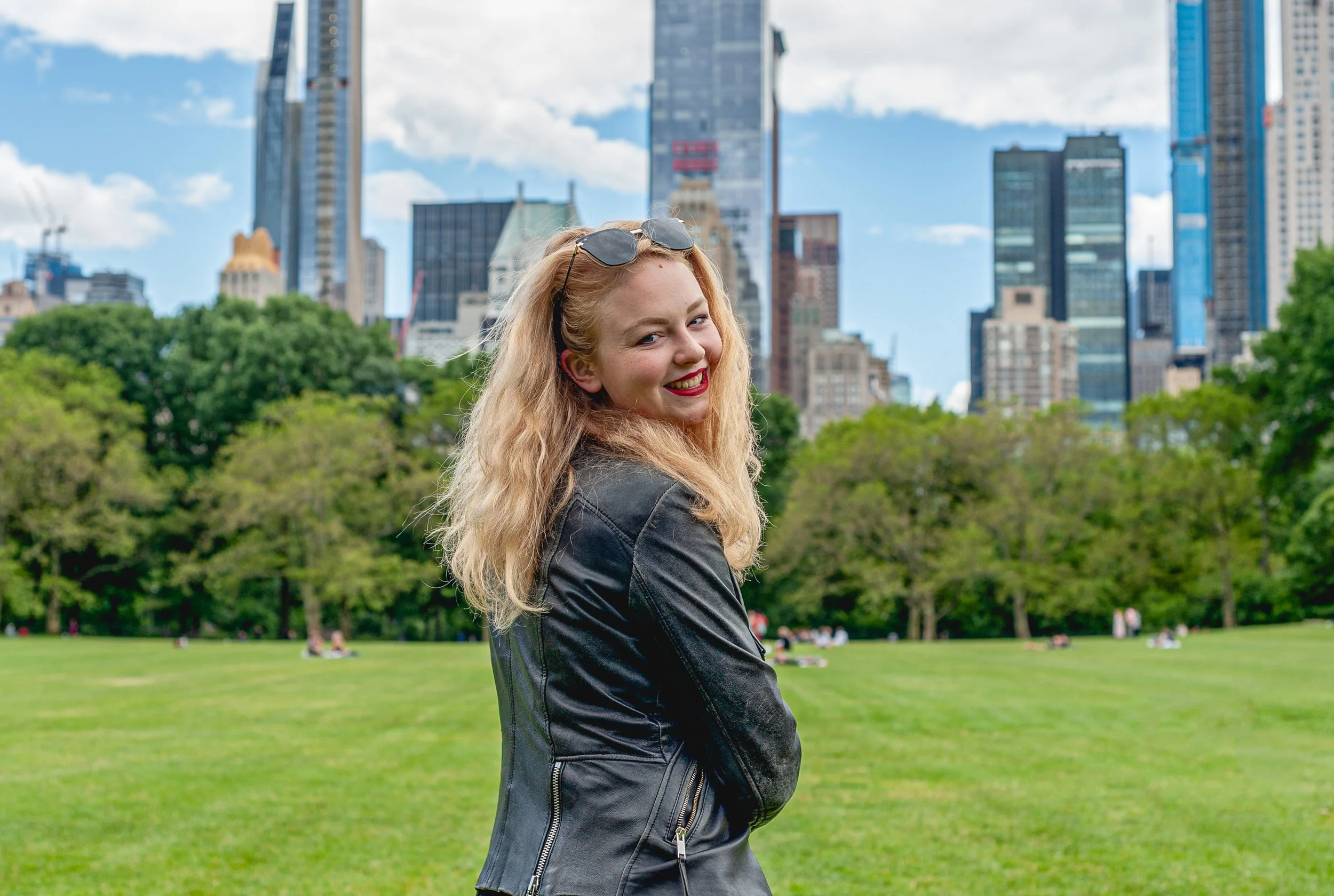 A smiling woman with blonde curly hair in a black leather jacket standing in a park with green grass and trees, city skyscrapers in the background.