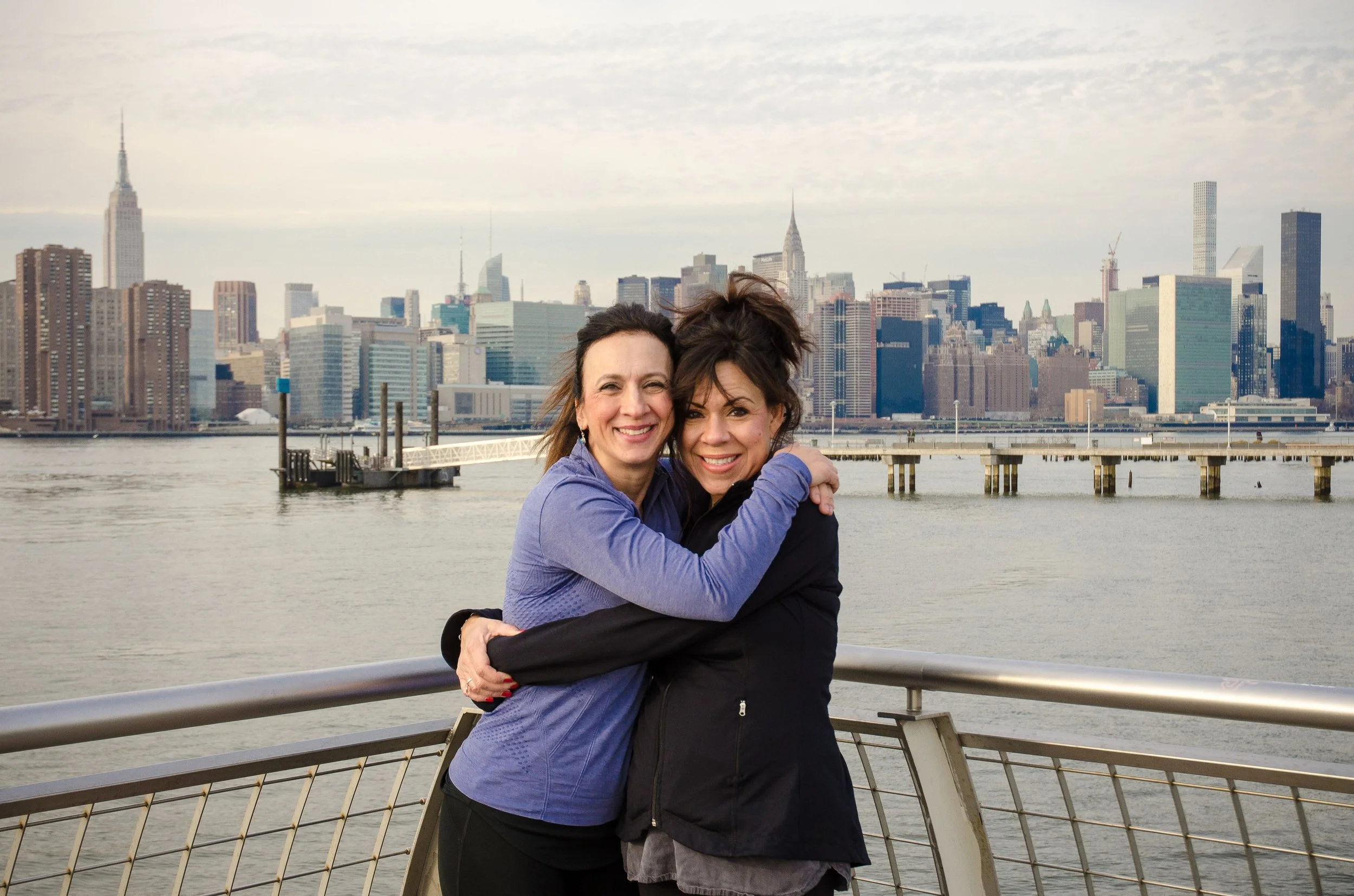 Two women hugging and smiling on a waterfront with New York City skyline in the background.