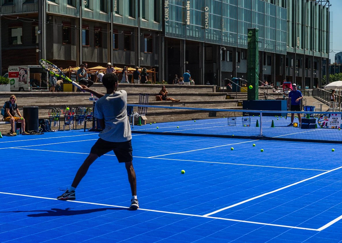 A young boy playing tennis on a bright blue outdoor tennis court in an urban area with a large glass building and spectators in the background.