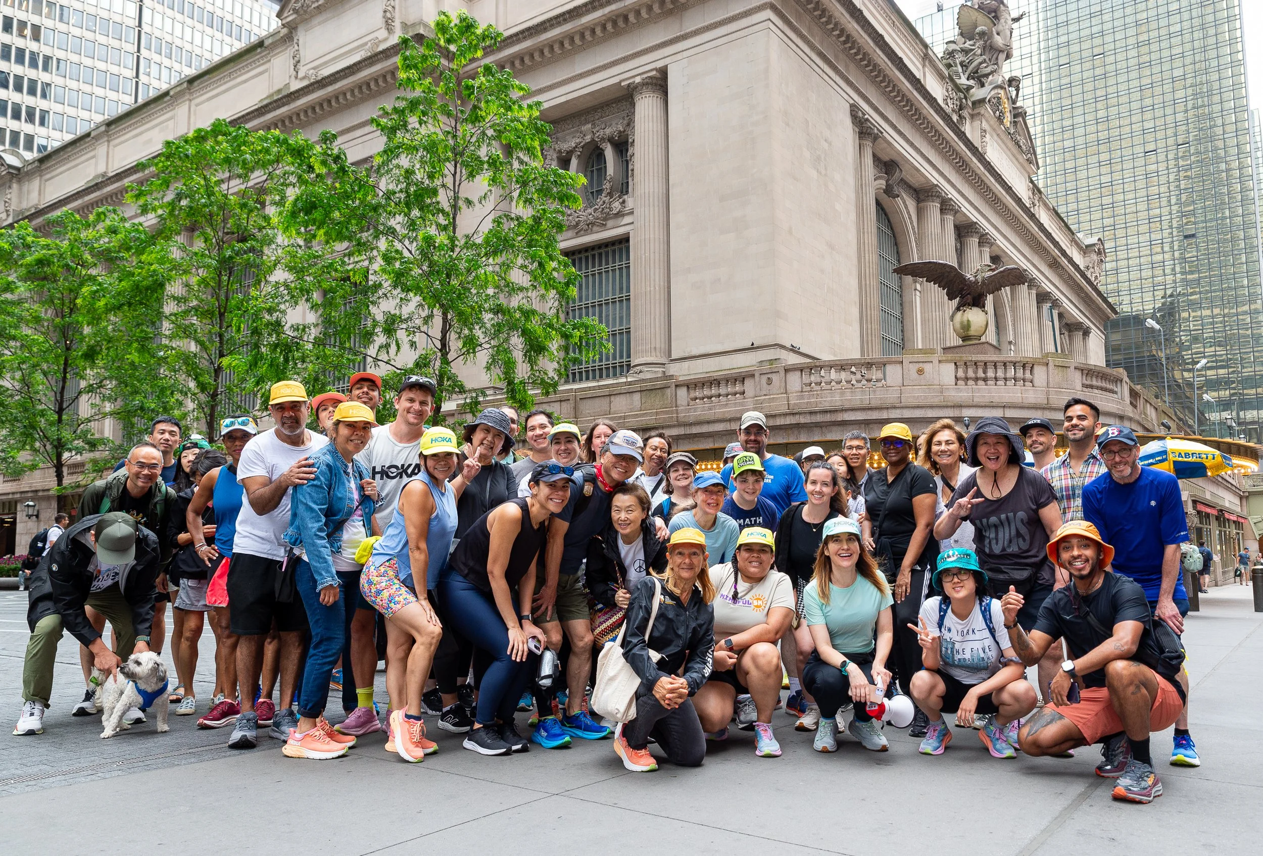 A large group of diverse people in athletic clothing and hats, posing happily for a photo in front of a historic building with columns and statues, surrounded by tall modern skyscrapers in an urban setting.
