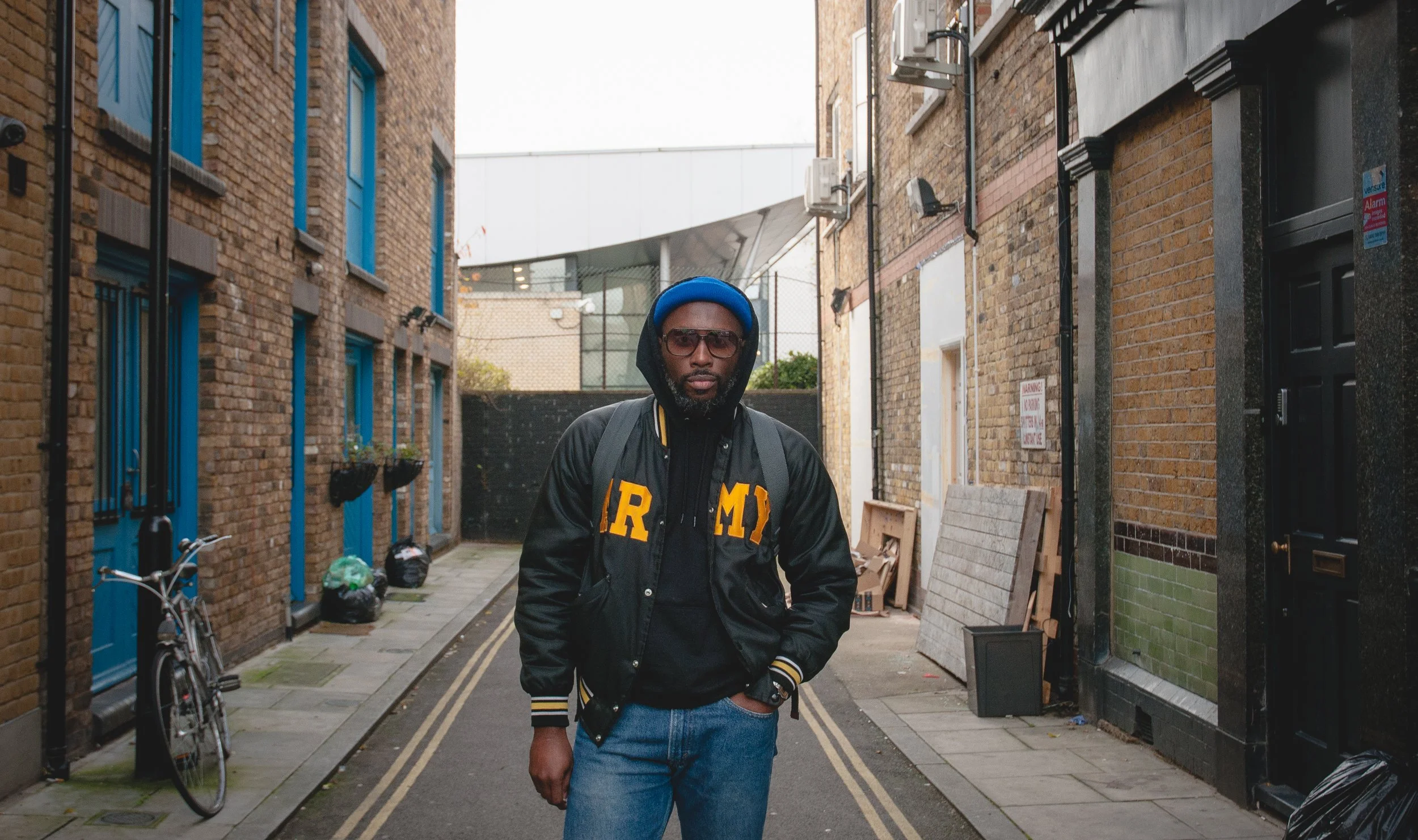 Street portrait of NYC photographer and director Marques Jackson wearing a black vintage Army jacket in an urban alley in Shoreditch, London.