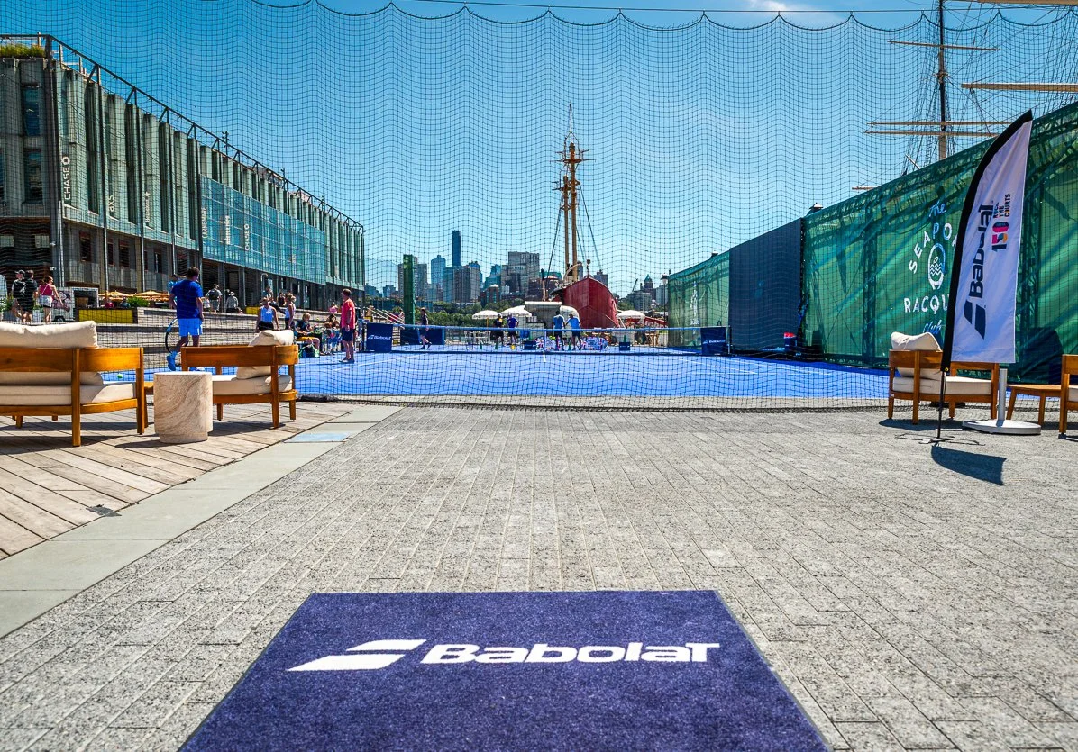A tennis court with a blue surface is set up outdoors in an urban area, enclosed by a net and green wind screens. There are people playing and standing around the court, with some in sports attire. In the foreground, there are chairs and seating area