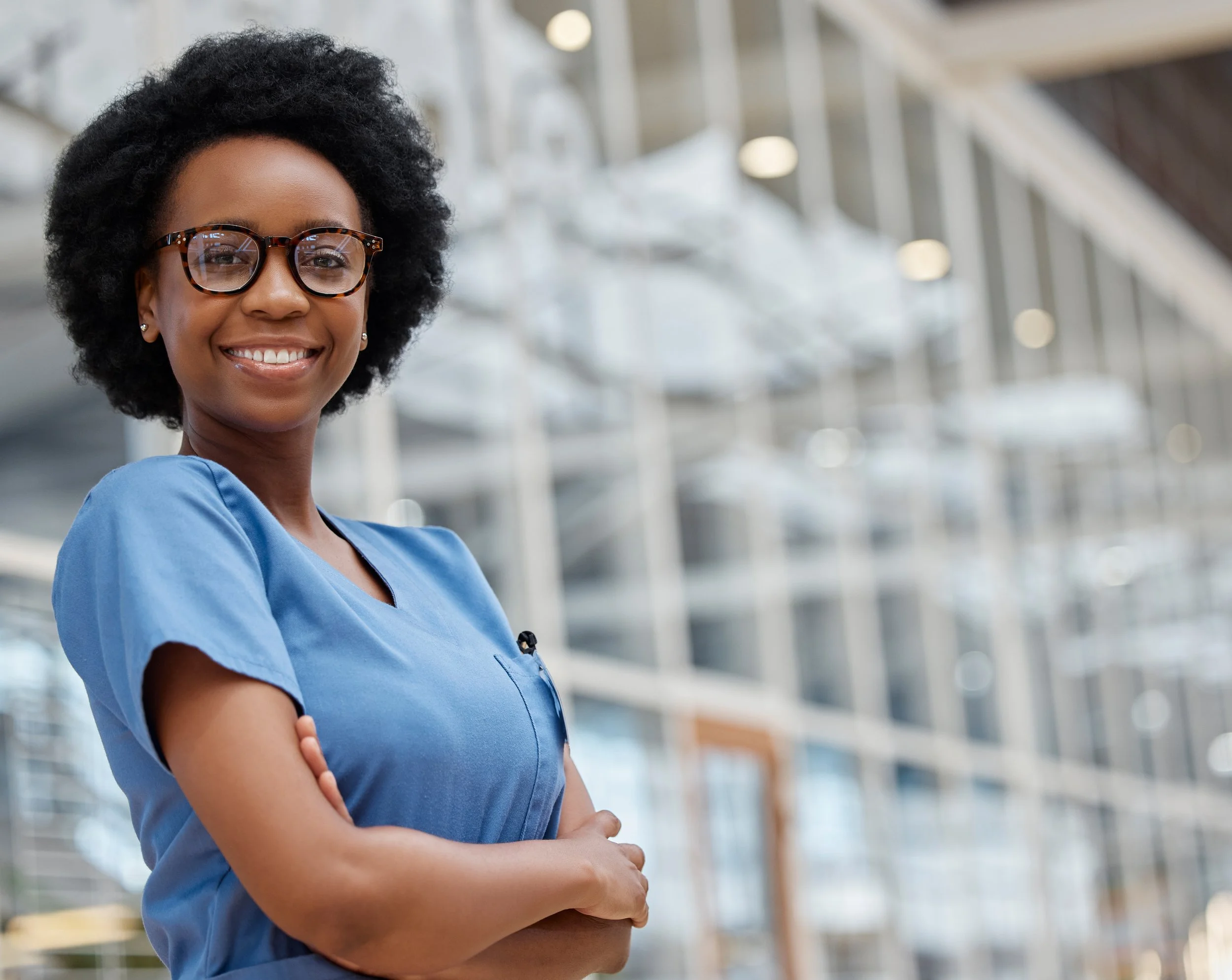 A female nurse is seen standing in blue scrubs in front of a hospital