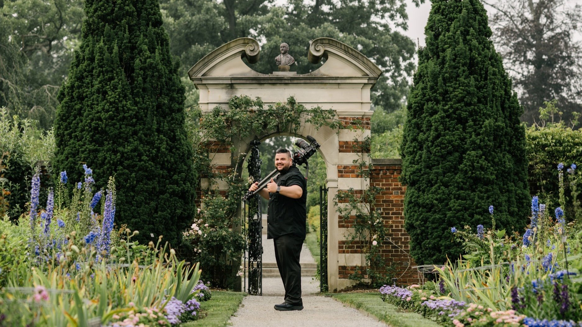 Man standing on garden pathway holding a camera crane, surrounded by colorful flowers and tall green trees, with an ornate stone archway in the background.