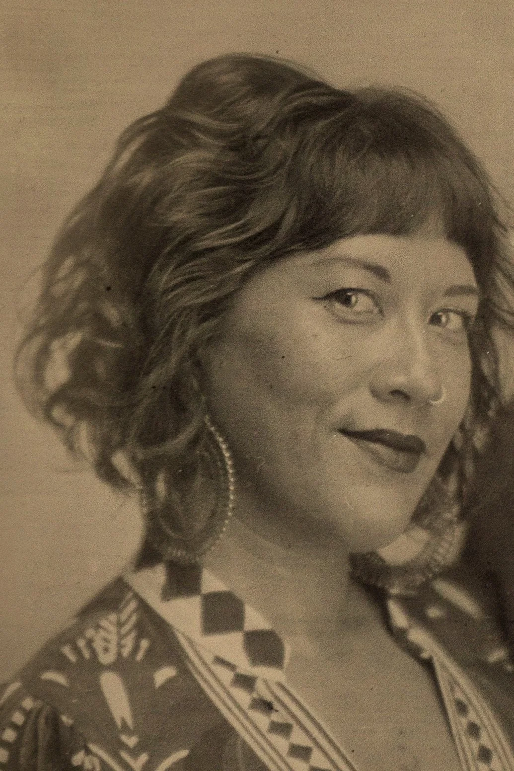 Black-and-white photograph of a woman with wavy hair, wearing earrings, makeup with eyeliner and lipstick, and a patterned garment.