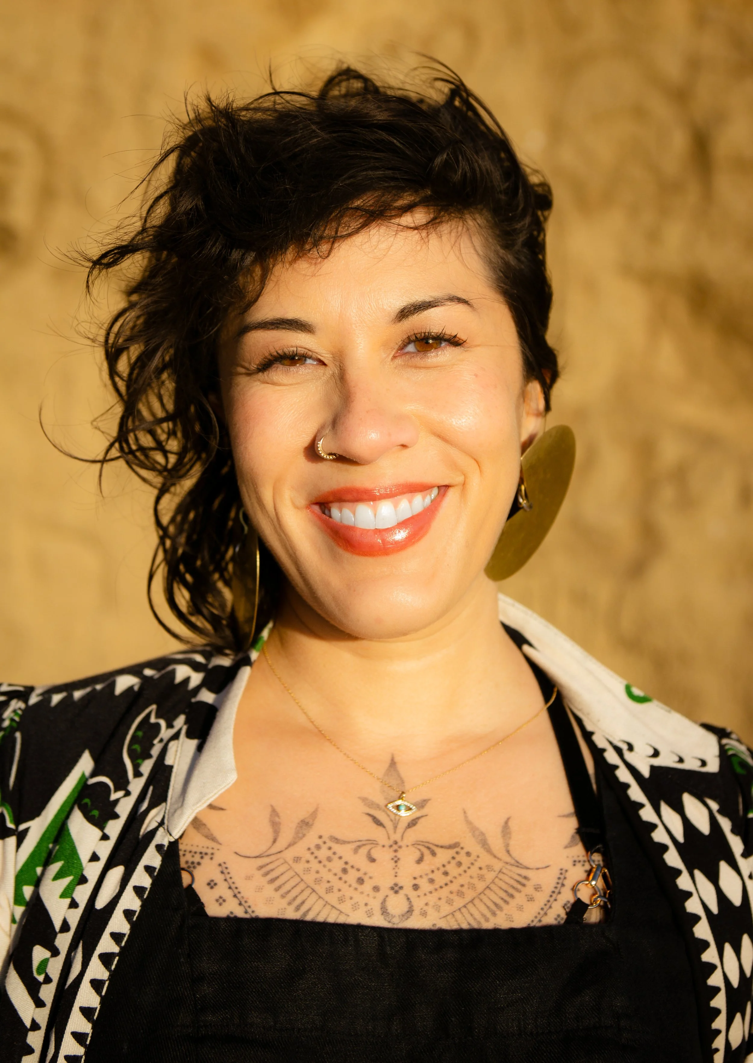 A smiling woman with short windswept dark brown hair, wearing gold hoop earrings, a nose ring, and a necklace with an eye-shaped pendant, standing against a textured gold, sandy wall on the beach.