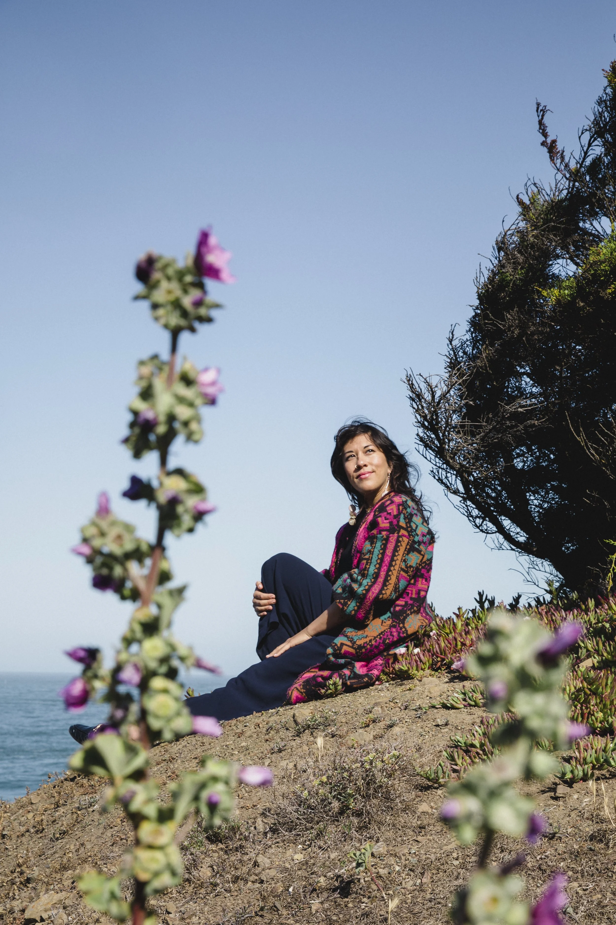 A woman in colorful clothing sitting on a hilltop with a scenic ocean view, wildflowers, and a clear blue sky in the background.