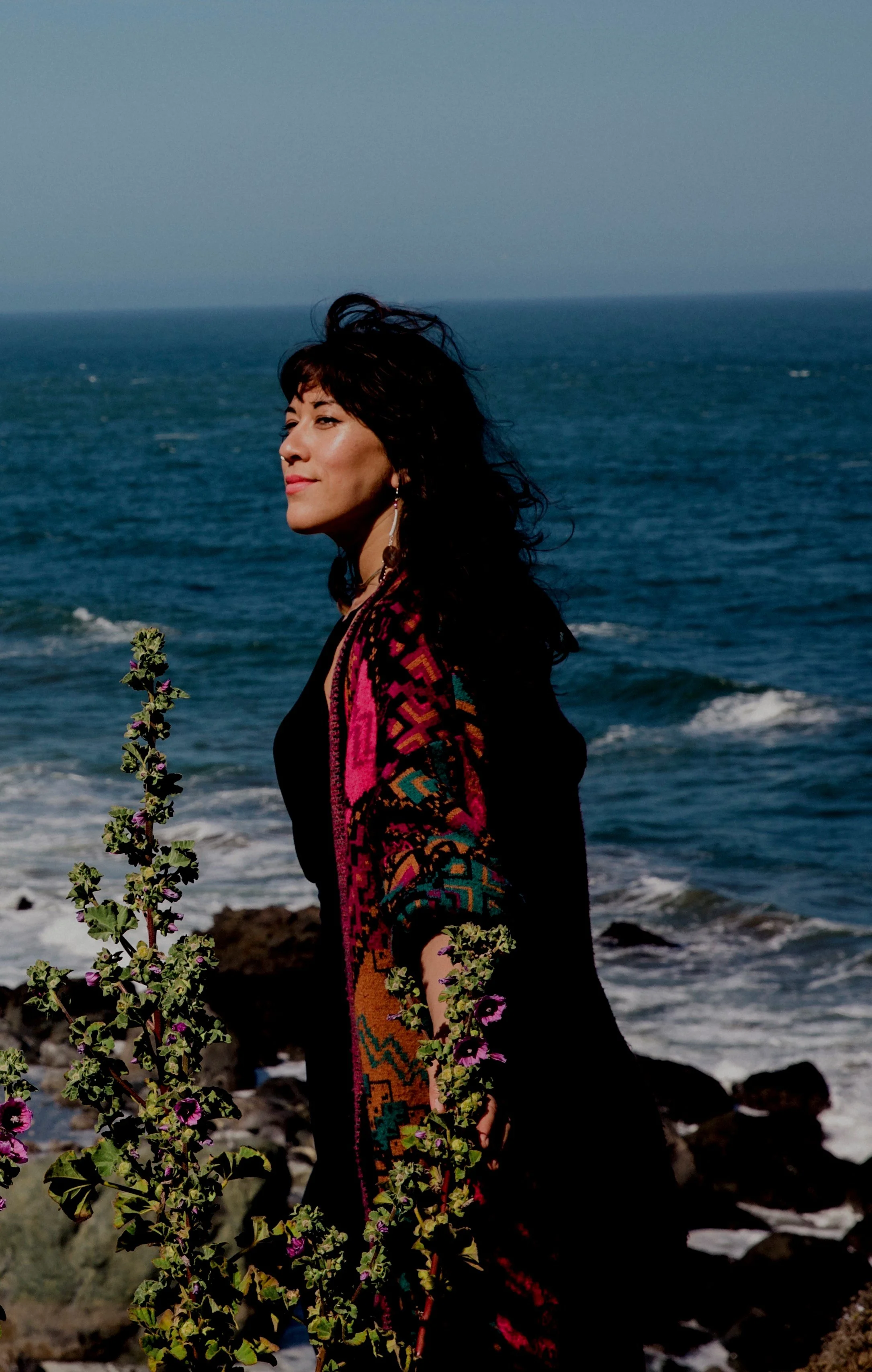 A woman with dark, wavy hair standing on the rocky shore, looking to the left with a calm expression, wearing a black outfit and a colorful, patterned shawl, with the ocean and clear sky in the background.