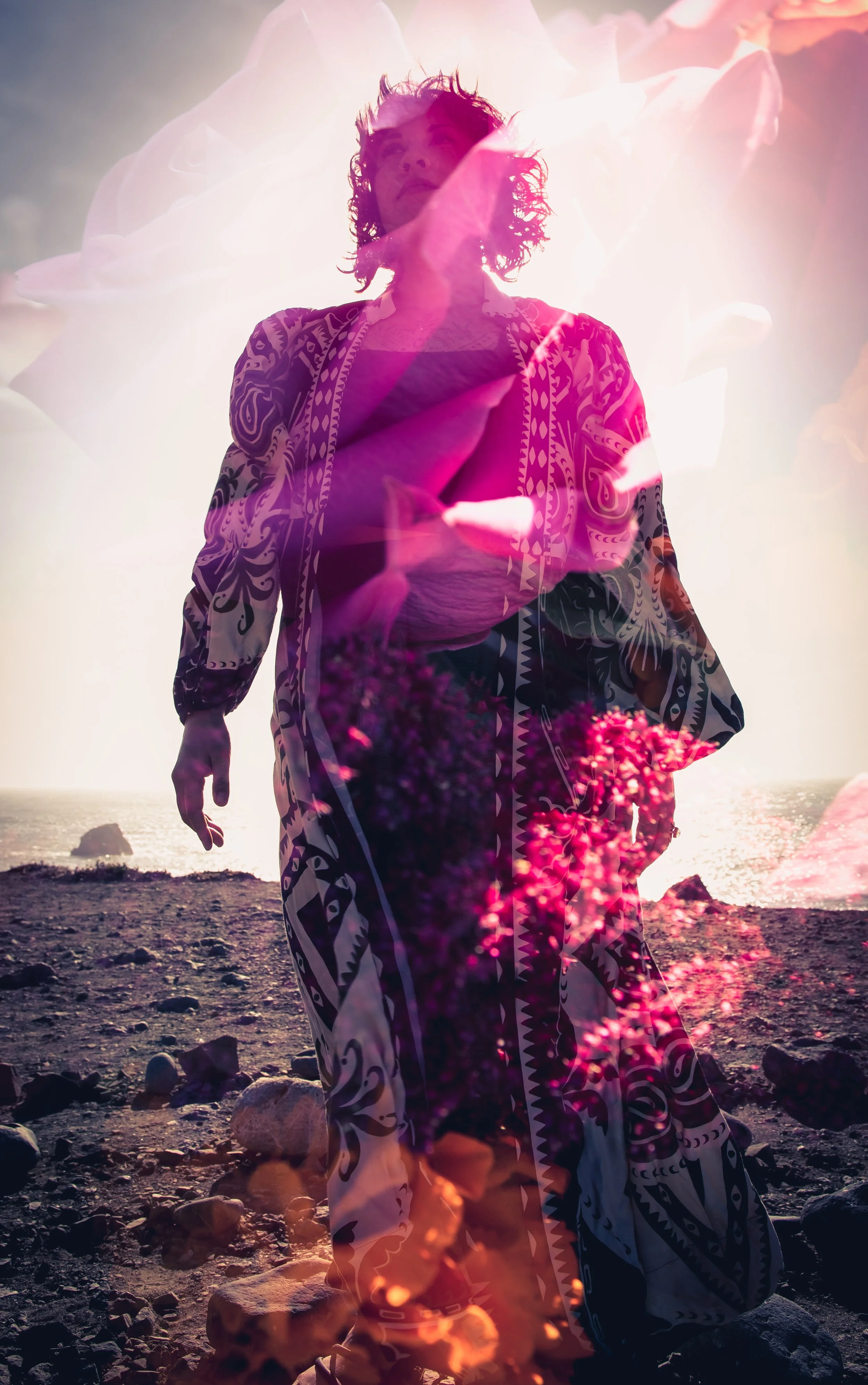 A woman with wavy hair walking on a beach at sunset, with pink flowers and soft sunlight creating an artistic, dreamy effect.
