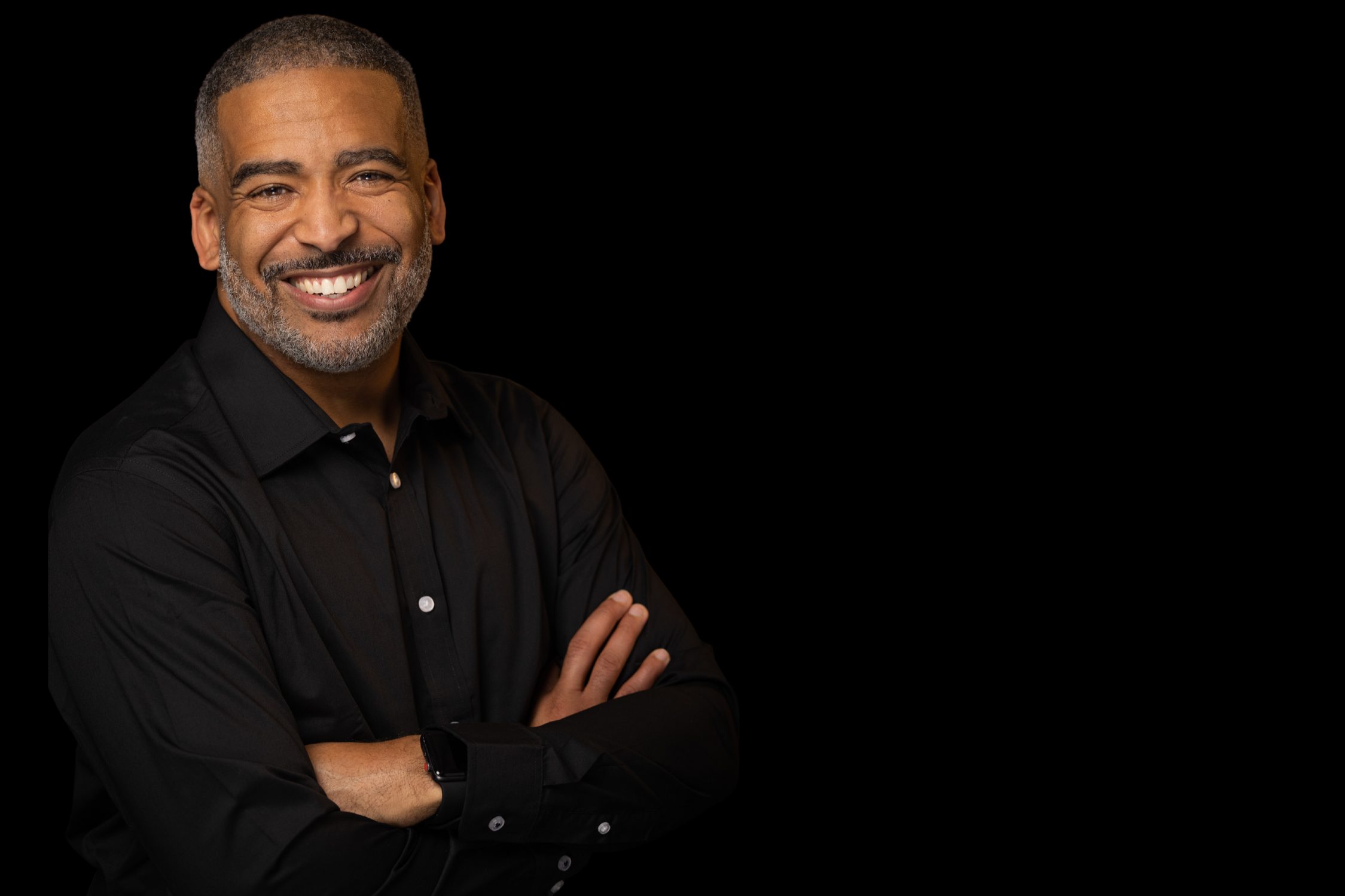 A smiling middle-aged man with short hair and a beard, wearing a black shirt with folded arms, against a plain black background.