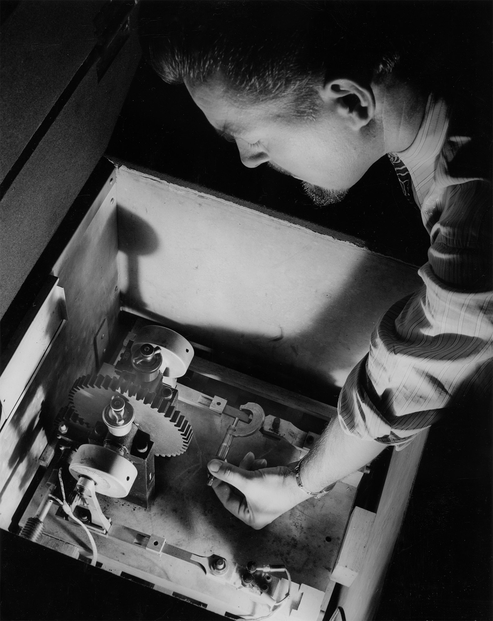 This combination oven-refrigerator that varies temperatures from 50 degrees below zero to 200 degrees above has been constructed at the Westinghouse Research Laboratories to test the strength of Micarta plastic used in making engine gears and aircraft parts. Frank Cassel, Westinghouse research engineer, is shown measuring the dimensions of a Micarta test piece in the oven-refrigerator before beginning a “fatigue” test. This Micarta stock, along with a similar one to its left, will be vibrated up and down 15 times every second, day and nights for as long as three months. While one end of the plastic stick is held stationary, the other end is moved up and down rapidly by the rotating arm and wheel. The amount of load applied and changes in the flexibility of the plastic are measured by the wire resistance “strain gauge” at the stationary end of the vibrator. Cooled by dry ice and heated by electric coils, the oven-refrigerator provides extremes of cold and heat to aid in the development of plastic equipment that gives maximum performance at any working temperature.
