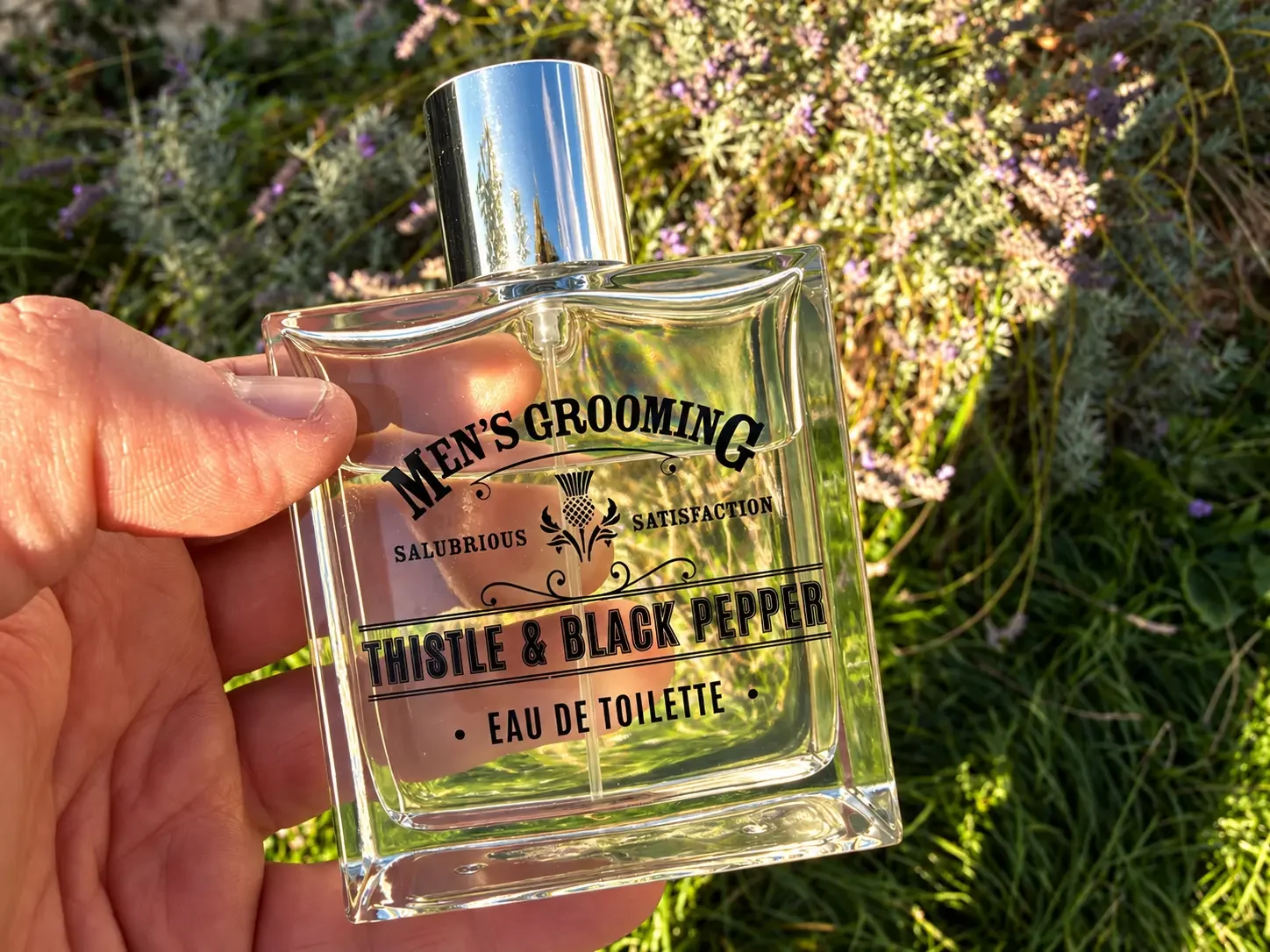 Men’s Grooming Thistle & Black Pepper Eau de Toilette bottle held by hand outdoors against a soft lavender and green garden background.