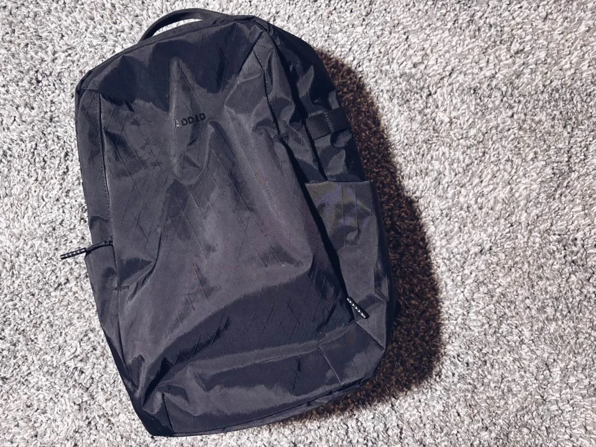 Front view of the ADD1D D2D Essential Backpack showing the minimalist shape and X-Pac fabric finish on a neutral background.