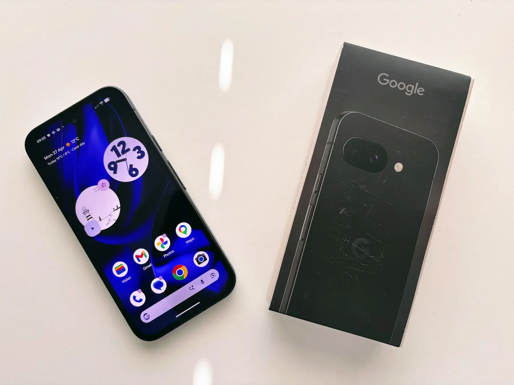 Top-down view of the Google Pixel 9A in Obsidian Black next to its retail box on a white desk