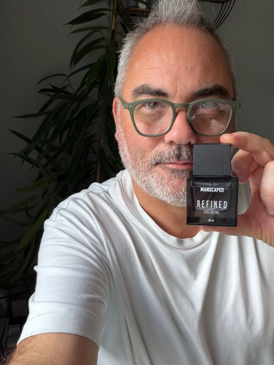 Man wearing green-framed glasses holding an uncapped bottle of Manscaped Refined cologne beside his face.