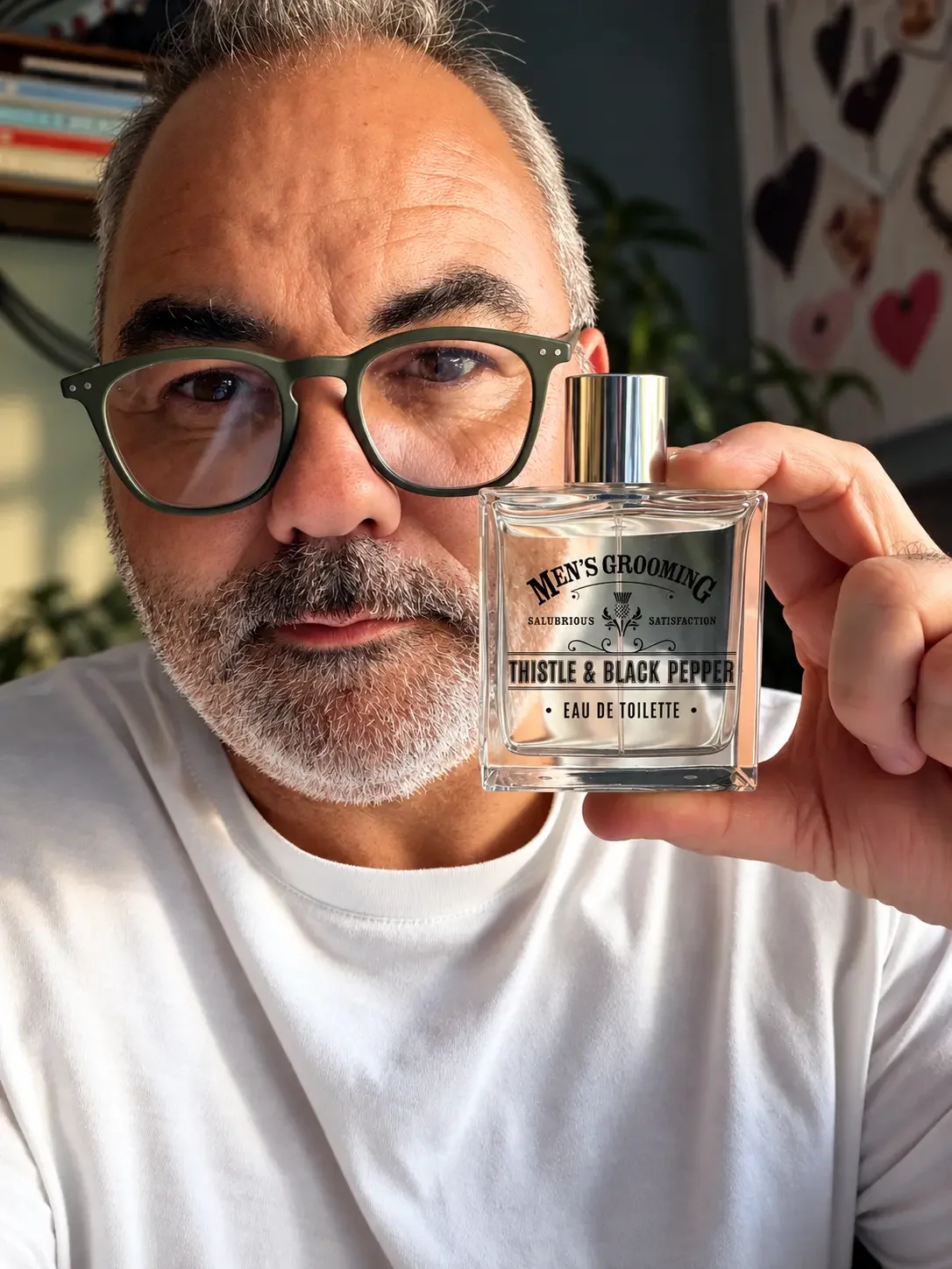 Portrait of a man holding Men’s Grooming Thistle & Black Pepper Eau de Toilette beside his face indoors in soft natural light.