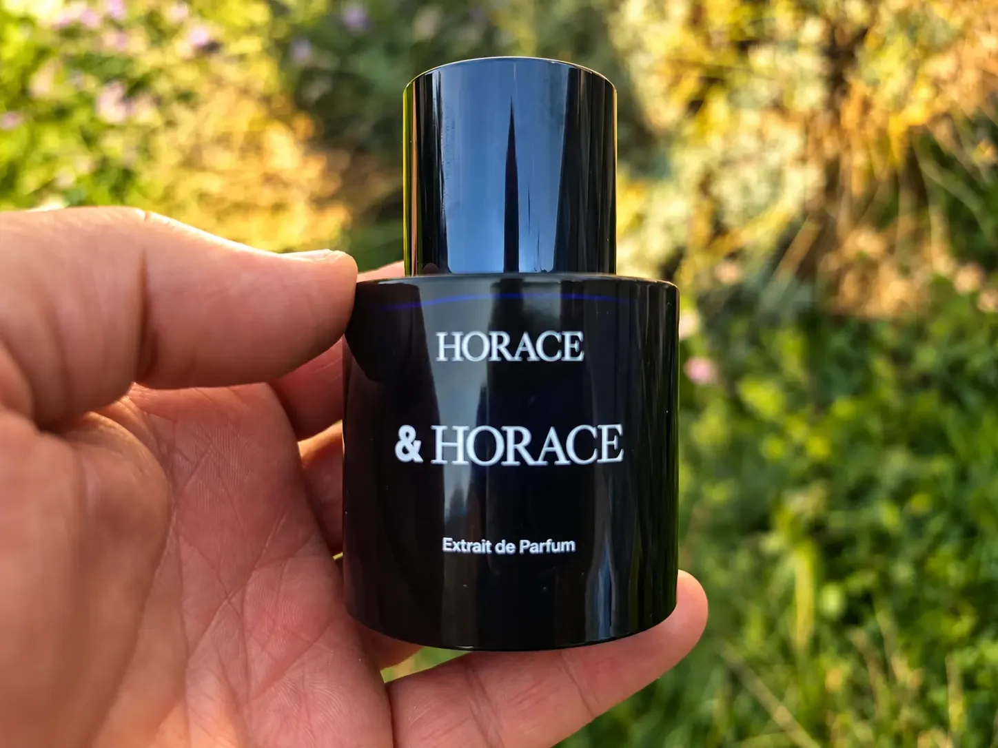 Horace Extrait de Parfum bottle held in hand outdoors with a soft green garden background.
