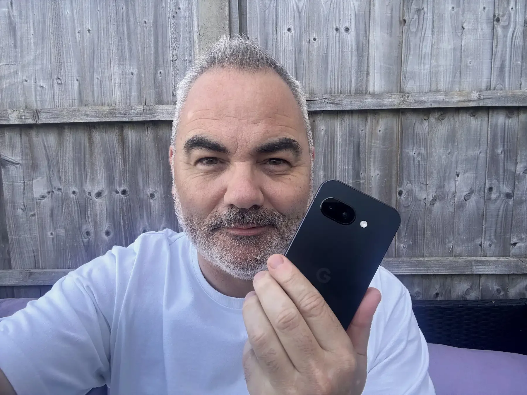 Jerome from Dapper & Groomed holding the Google Pixel 9A in front of a wooden fence, demonstrating the phone's size and design.