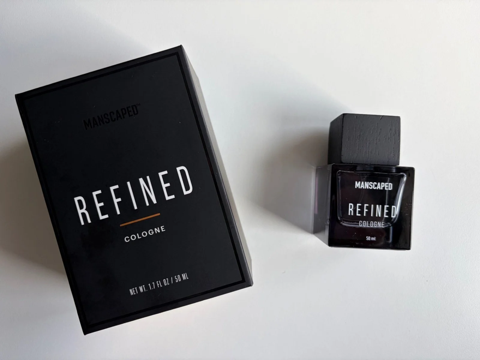 Manscaped Refined 50ml cologne bottle beside its black presentation box on a white surface.