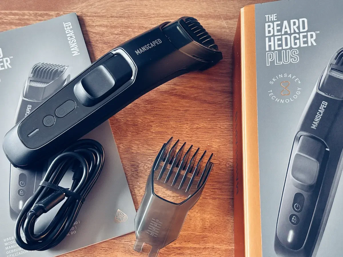 Manscaped The Beard Hedger Plus trimmer with box, comb attachment, USB-C cable and instructions on a wooden table.