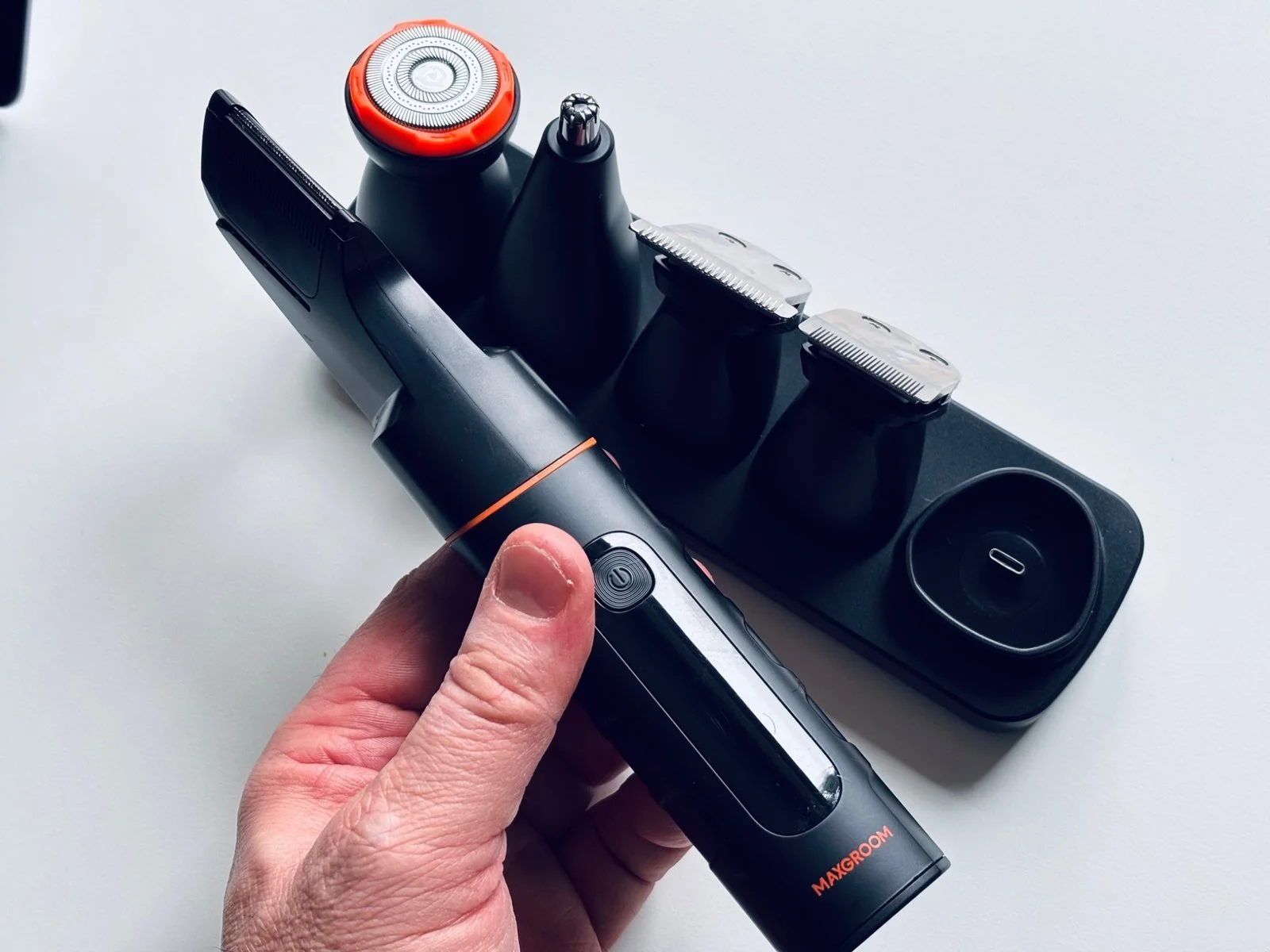 Handheld view of the Maxgroom all-in-one body hair trimmer showing its ergonomic grip, modern design and useful charging dock with multiple heads.