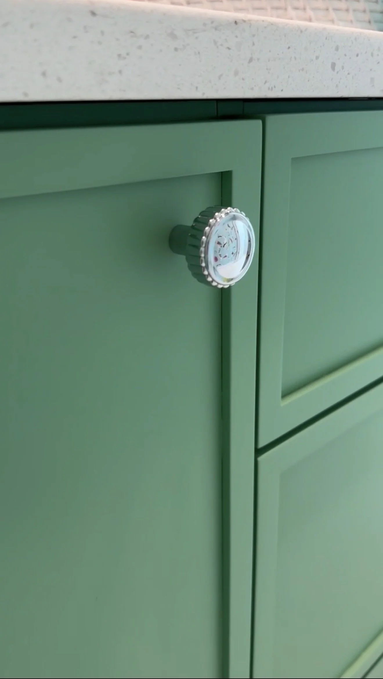 Close-up of a green cabinet with a textured white countertop, showing a round, silver-colored knob on the door.