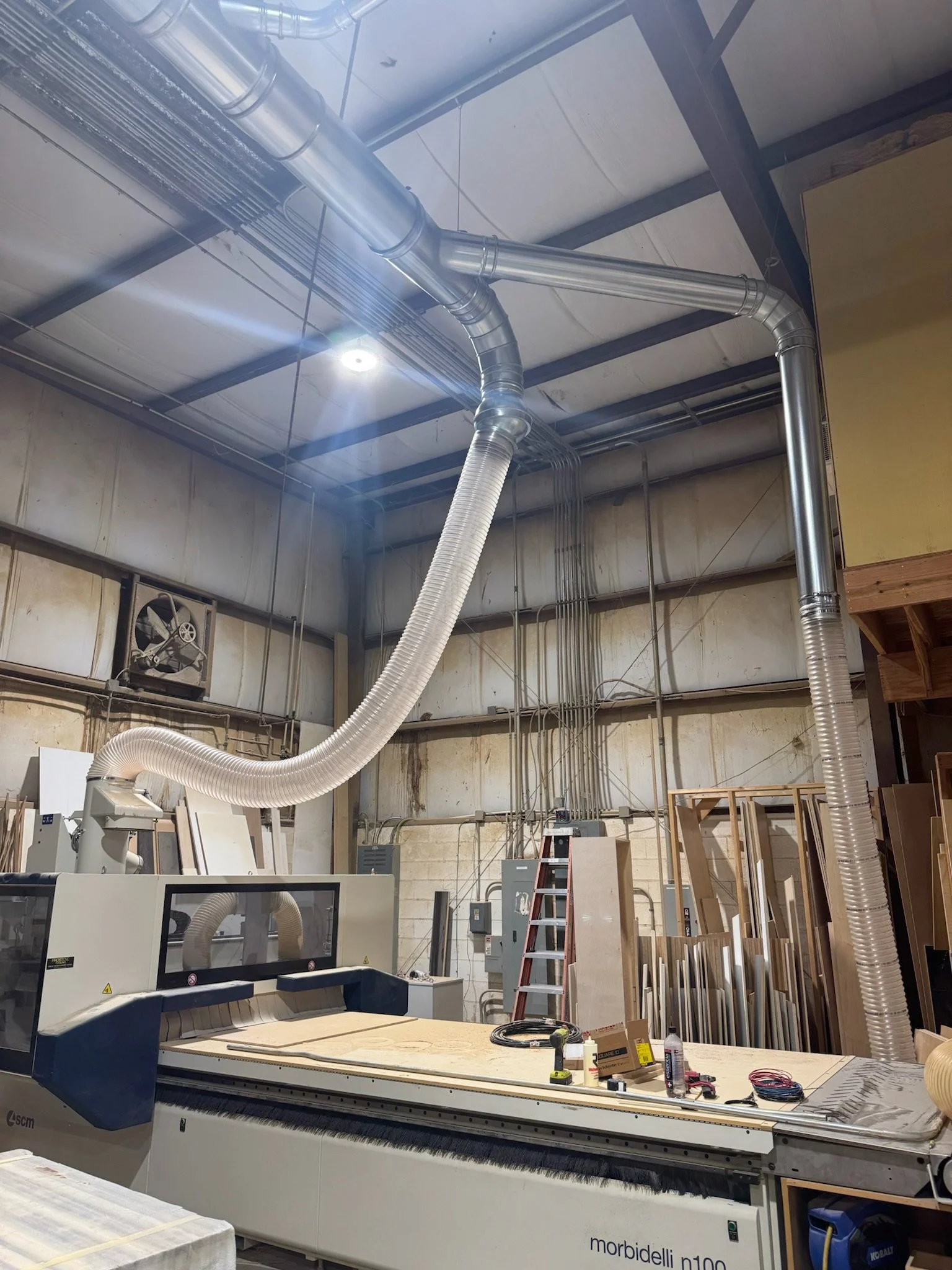 Workshop or manufacturing space with large CNC machine, wood pieces, ladder, and industrial ventilation pipes.