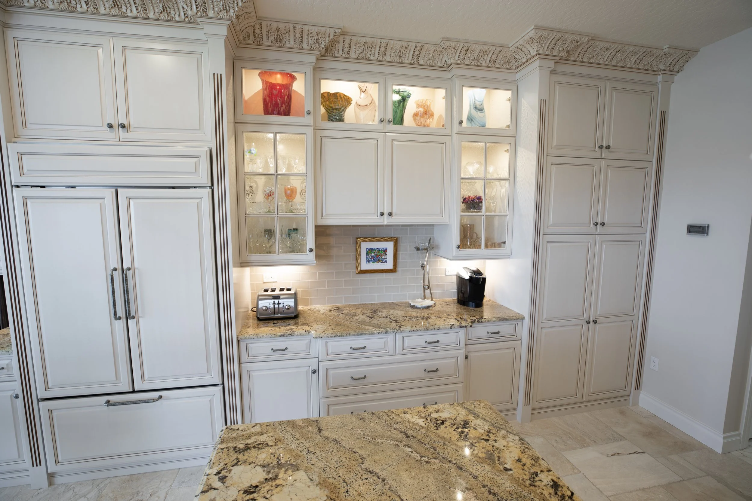 Kitchen with white cabinets, glass display cabinets with colorful vases, granite countertop, small kitchen appliances, framed artwork, and beige tiled floor.