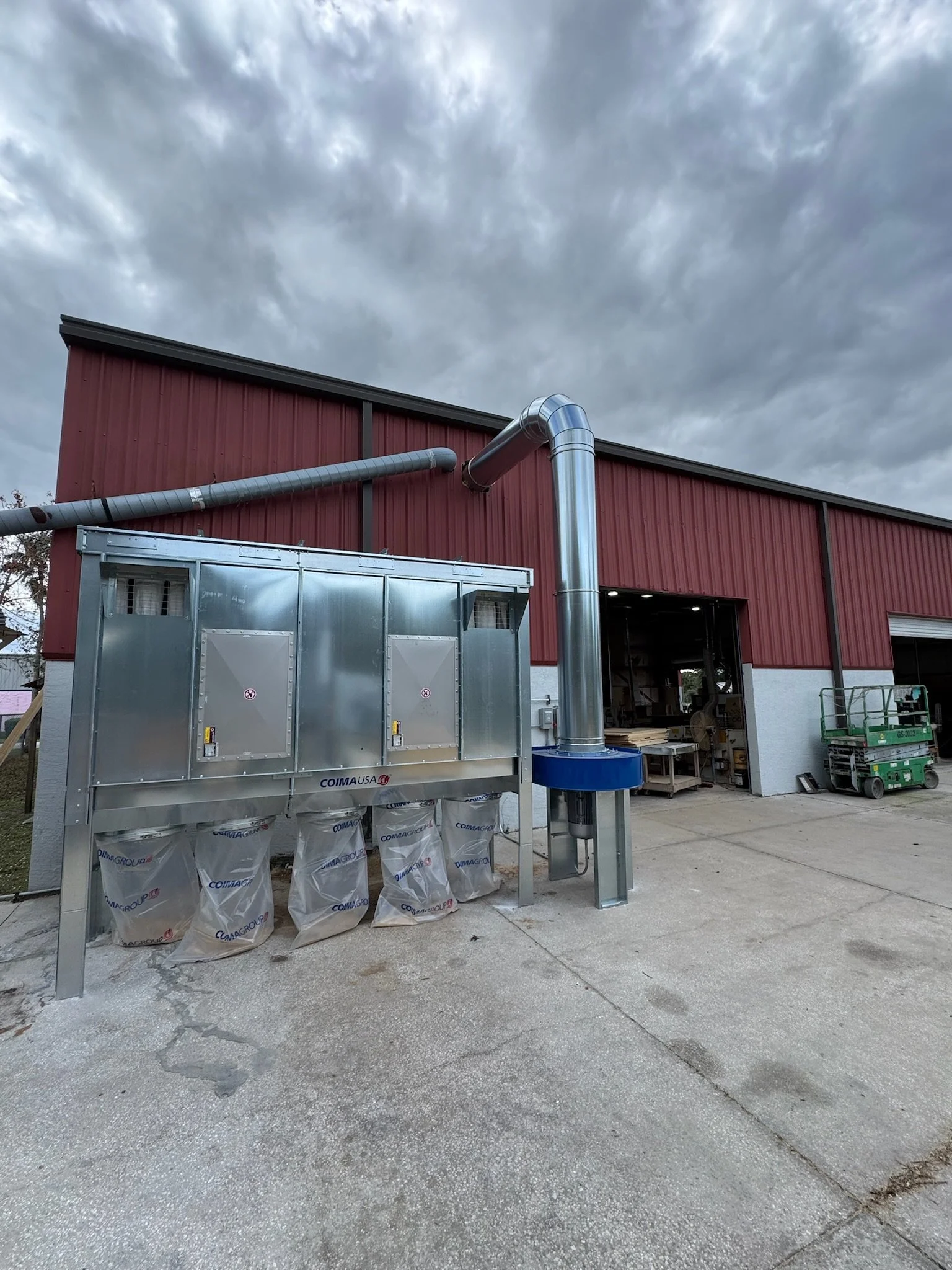 Exterior view of a red metal building with industrial ventilation equipment and pipes, surrounded by a concrete ground and overcast gray sky.