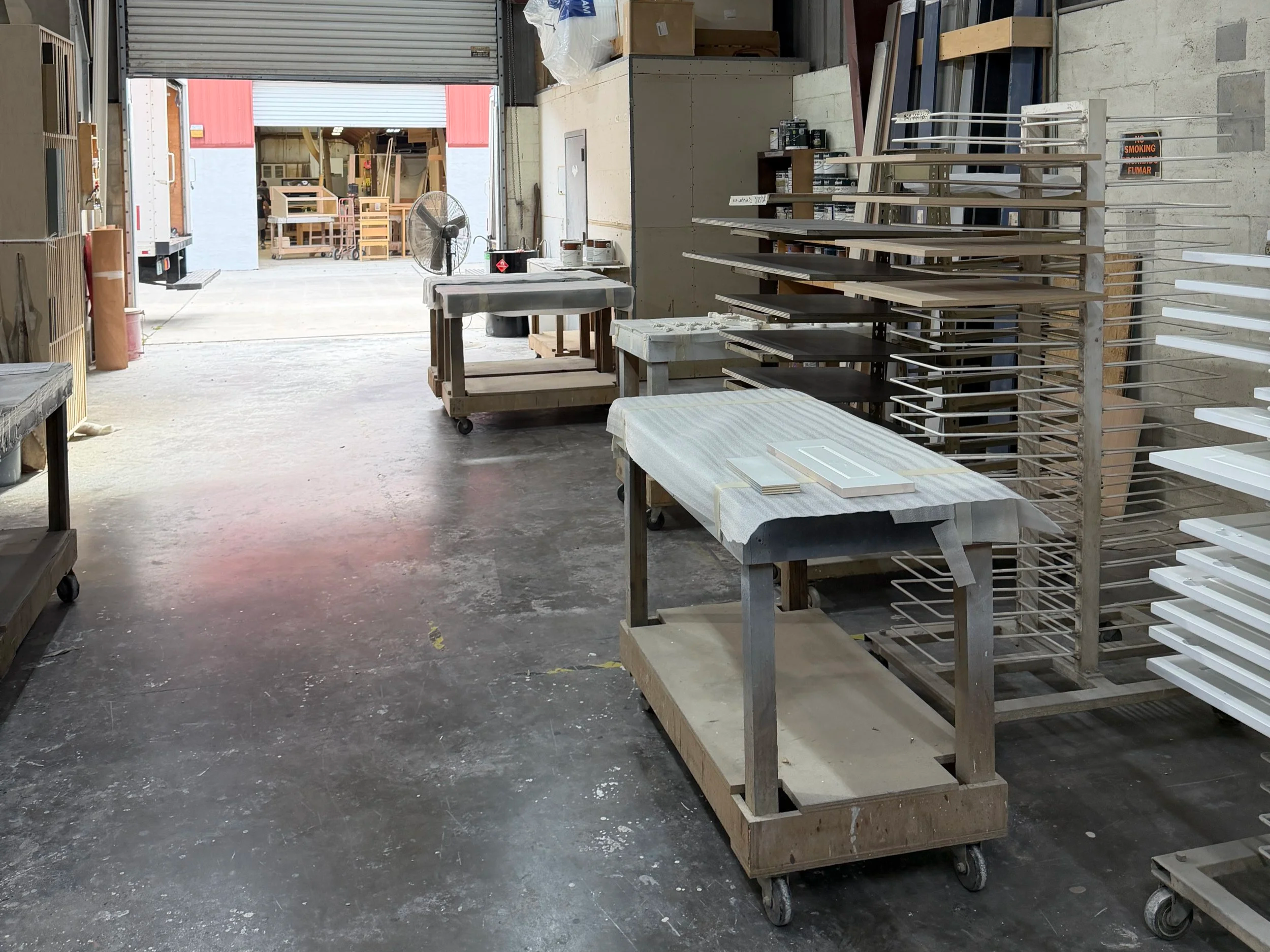 The image shows the interior of a warehouse with metal shelves and carts, some covered with fabrics. There are various materials and tools around, with an open large garage door leading outside to other stored items and equipment.
