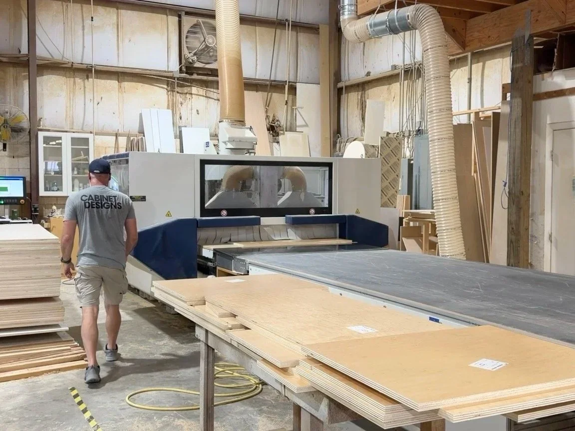 A man working in a woodworking shop with a large CNC machine cutting wood panels, stacks of plywood on nearby tables, and wood shavings on the floor.