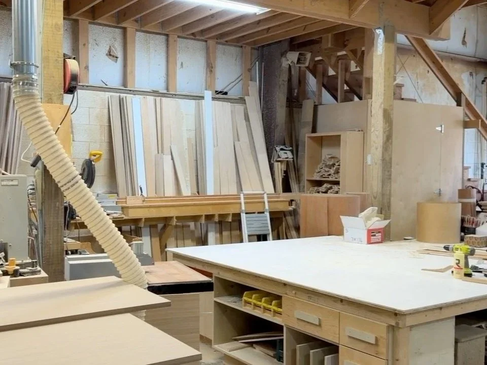 A woodworking workshop with various wood planks, tools, and equipment, including a workbench, storage shelves, and a dust collection hose.