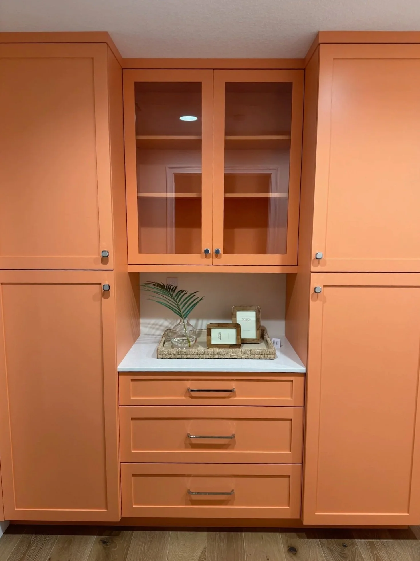 Wooden kitchen cabinet with glass doors, a white countertop, decorative tray and picture frames, and a small green plant.