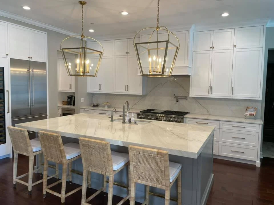 Modern kitchen with white cabinets, marble countertop island, and pendant lights hanging above the island. Four wicker bar stools are at the island, and stainless steel appliances are visible.
