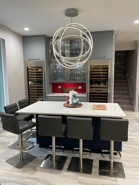 Modern kitchen with a white island, black bar stools, gray cabinetry, a red backsplash, built-in wine refrigerators, and a unique circular pendant light fixture.