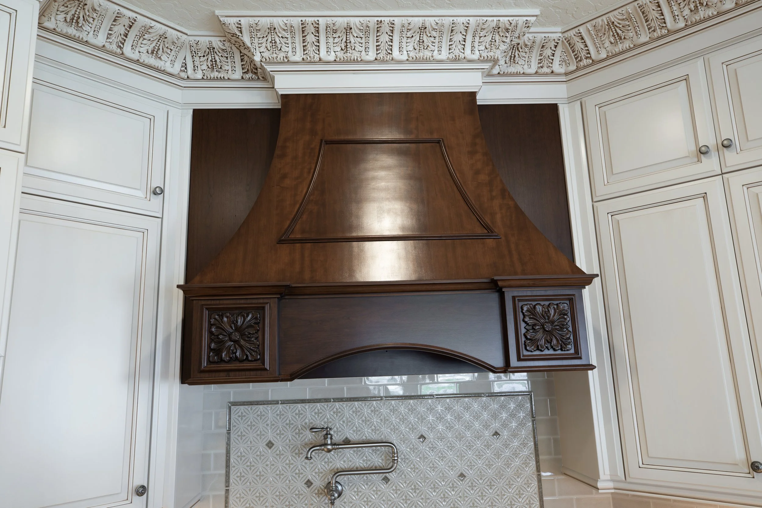 A vintage kitchen wood range hood with ornate carvings, surrounded by cream-colored cabinets with silver knobs, above a tiled backsplash with a floral patterned texture.