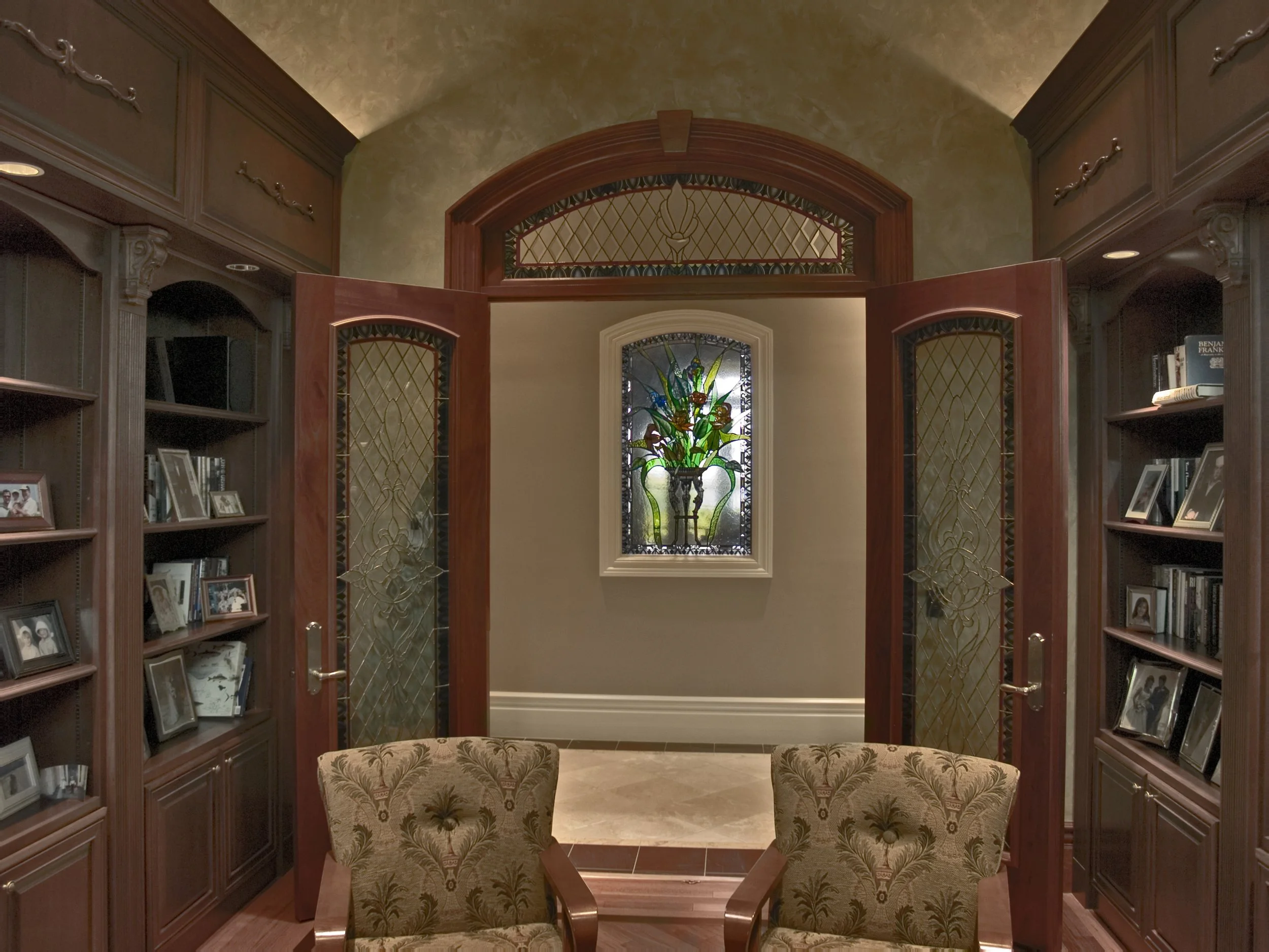 A room with wooden bookshelves on either side, a stained glass window with floral design, and double doors with stained glass accents. Two patterned chairs face the doors.