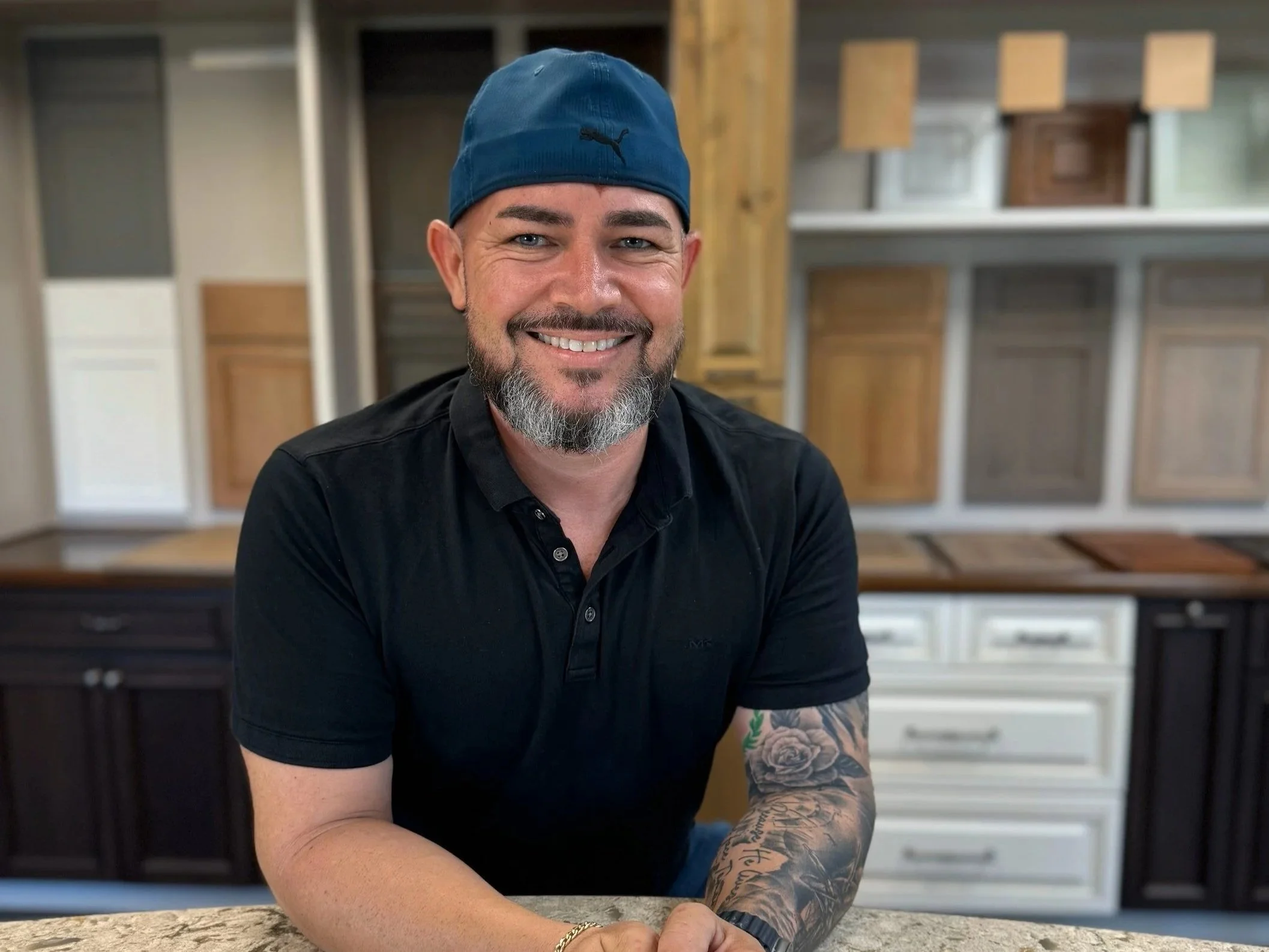 A man with a blue cap, dark shirt, beard, and tattoos on his arm smiling while sitting at a table in a woodworking or furniture store with wood samples and cabinet doors displayed in the background.