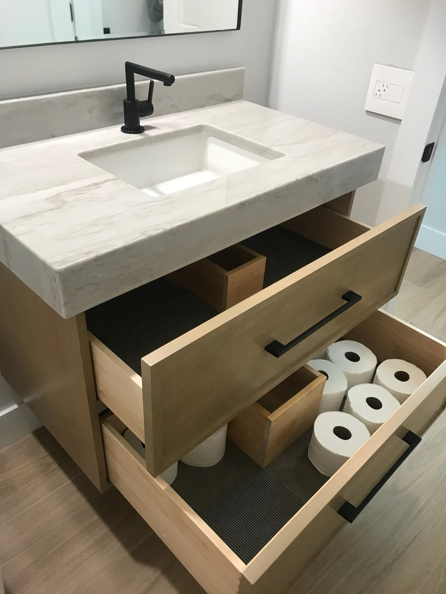 Open drawer vanity cabinet with a marble countertop, black faucet, and toilet paper rolls underneath.
