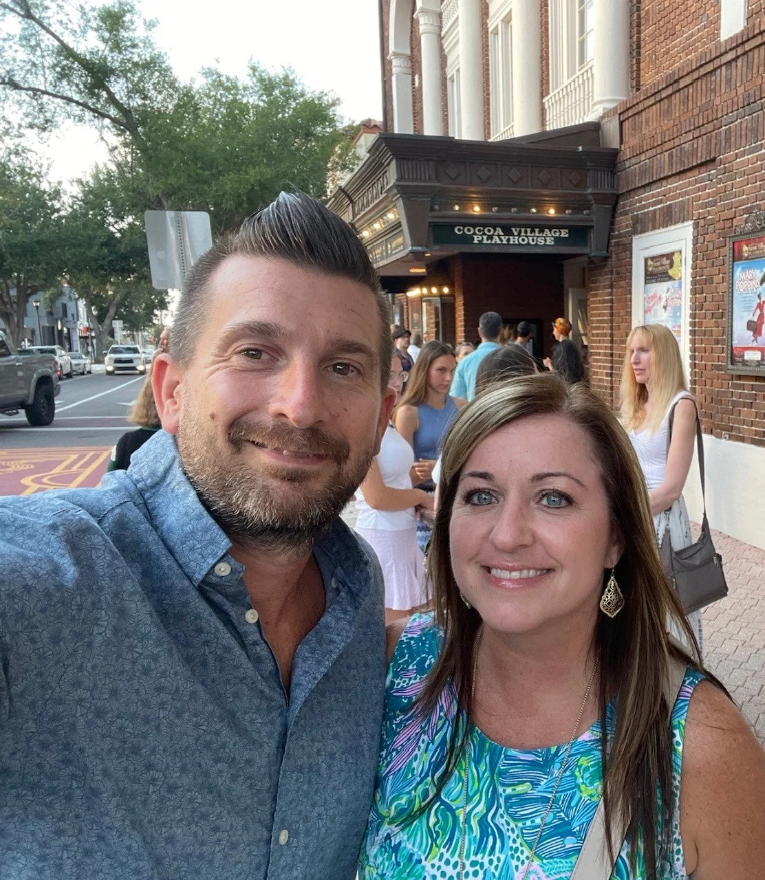 Two smiling people, a man with dark hair and beard in a blue shirt, and a woman with long brown hair in a colorful dress, taking a selfie outside the Cocoa Village Playhouse, with a line of people and a brick building behind them.
