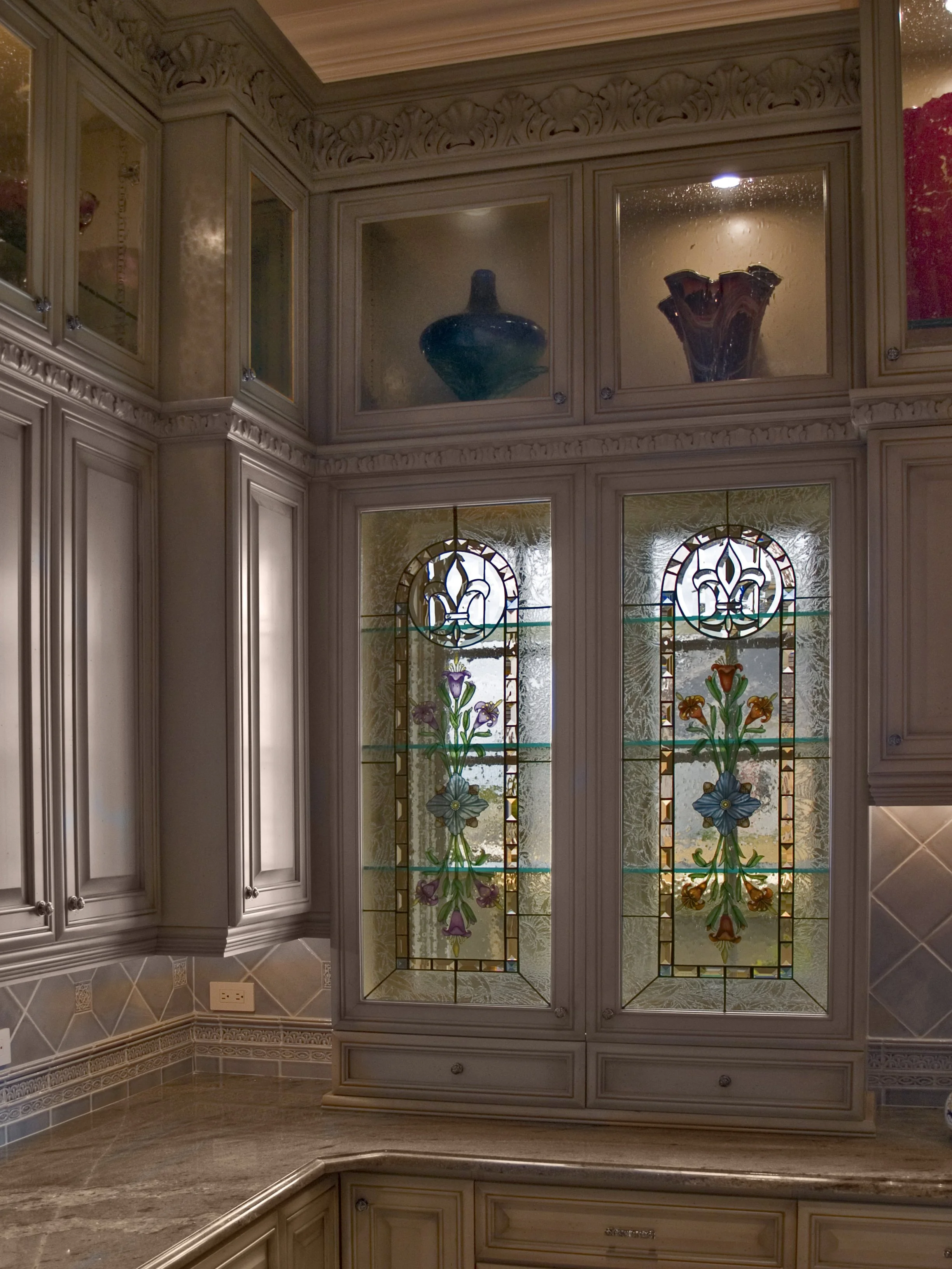A kitchen corner with beige cabinetry, stained glass panels with floral designs, and a marble countertop.