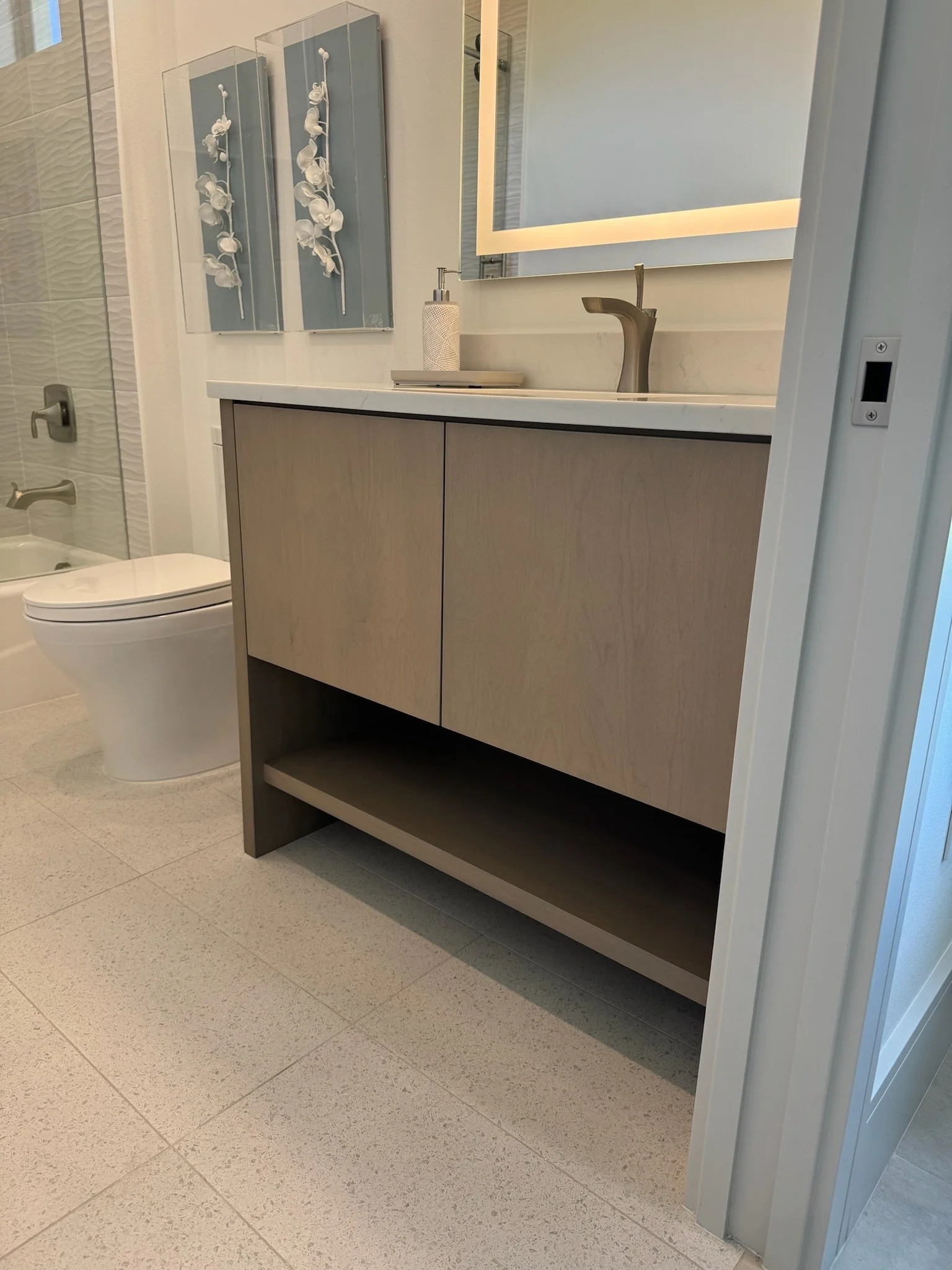A modern bathroom with a light wood vanity, marble countertop, and a mirror with backlit LED lighting. To the left, there's a toilet and a walk-in shower with textured white tiles.