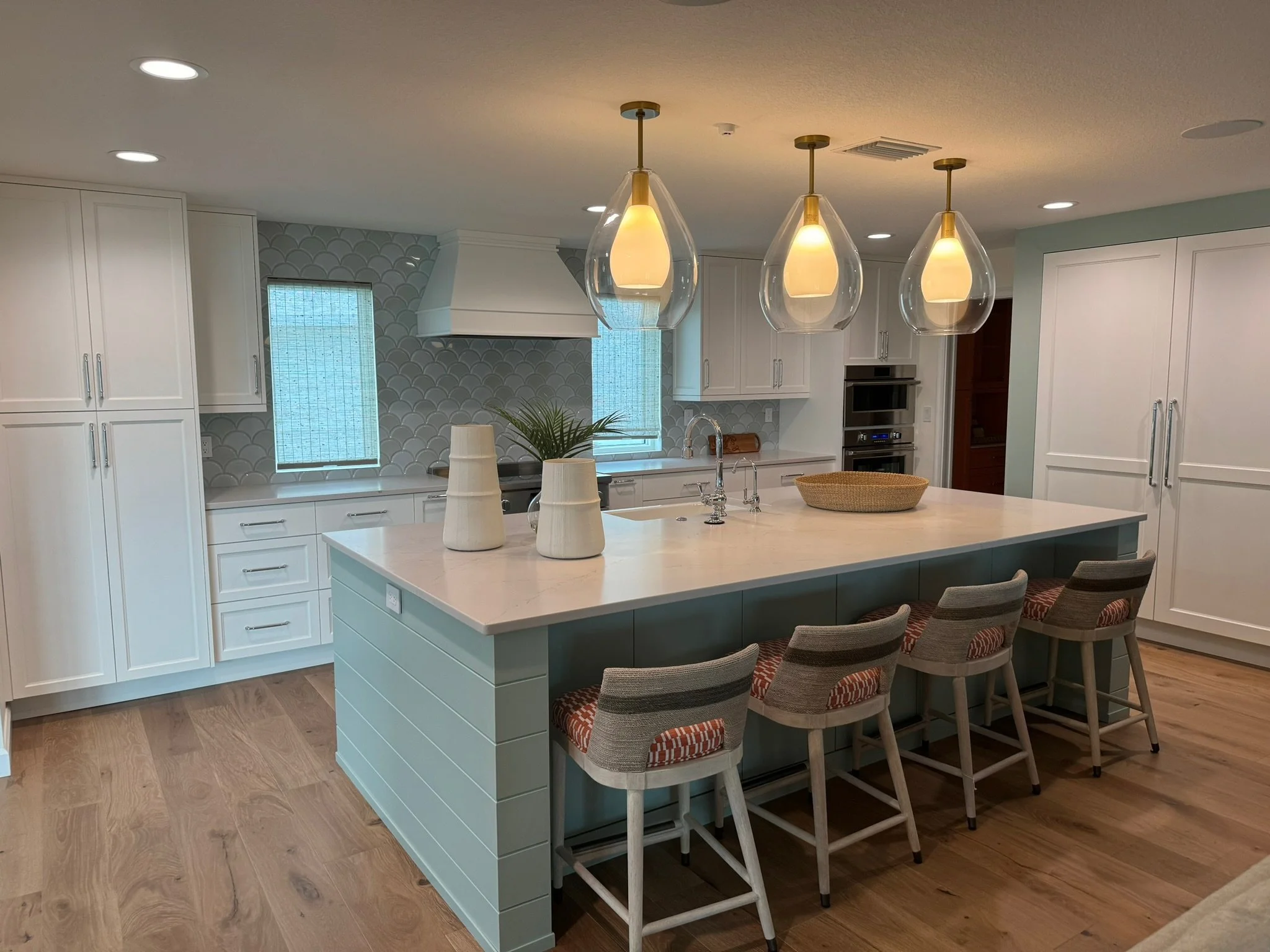 Modern kitchen with white cabinets, a large island with a light-colored countertop, and three teardrop-shaped pendant lights hanging above. There are two windows with blinds, a potted plant, and dining chairs with orange and gray patterned cushions a