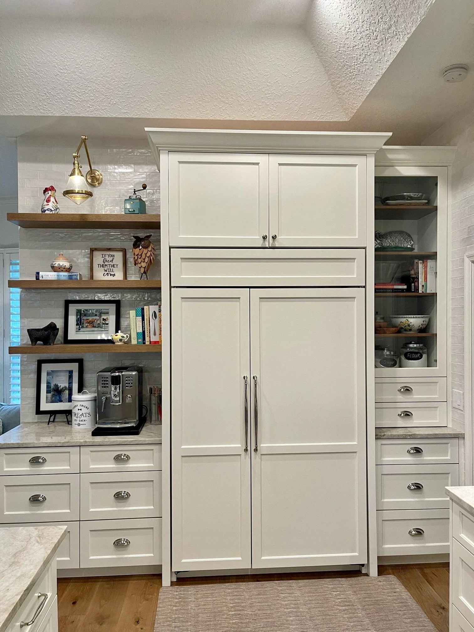 The image shows a white kitchen cabinet with multiple drawers and doors, surrounded by open shelves with decorative items, books, and kitchenware, on a wooden floor with a beige rug in front.