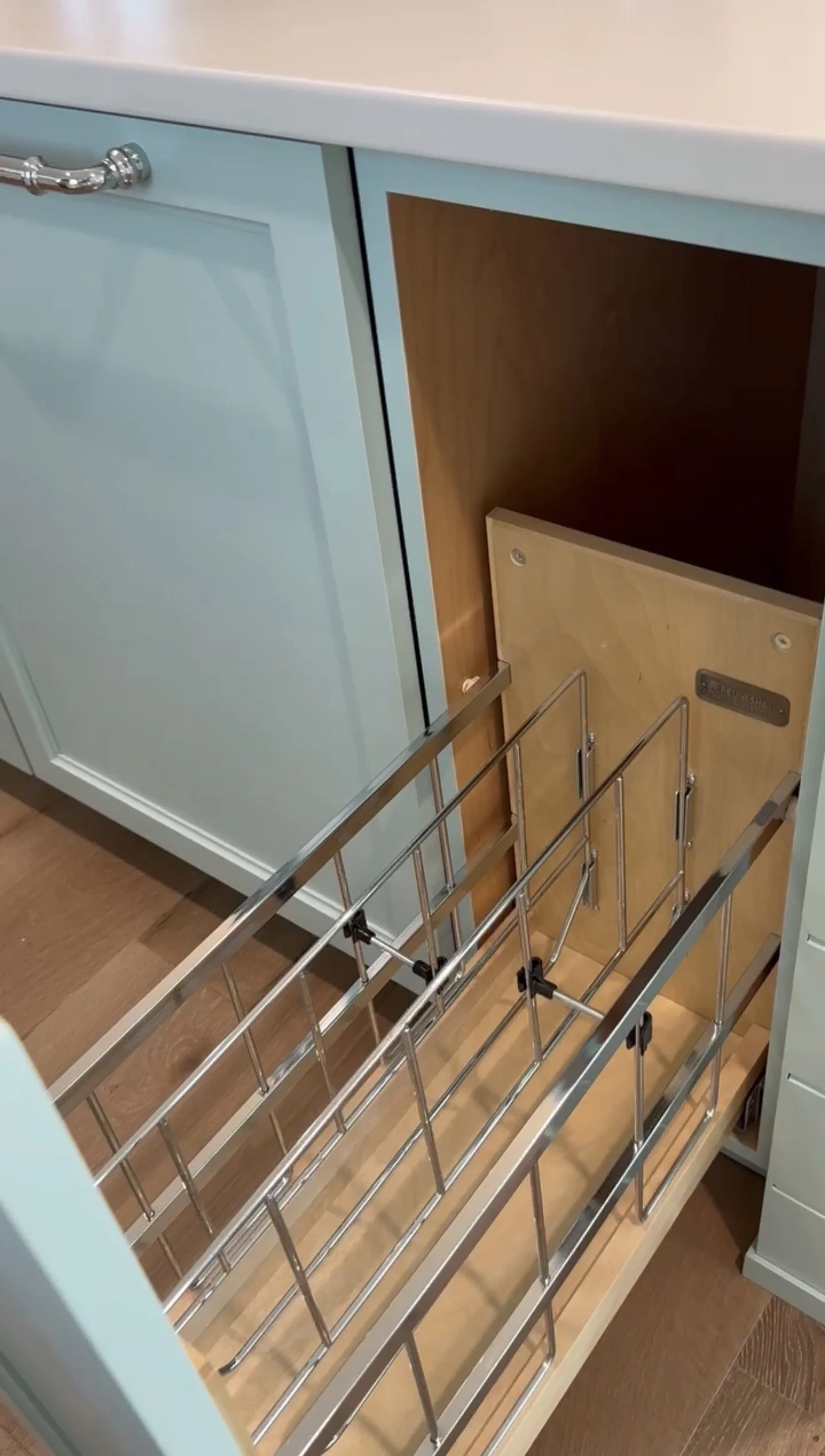 Open kitchen lower cabinet drawer with metal wire baskets and a wooden interior, with a pale blue front and a white countertop.