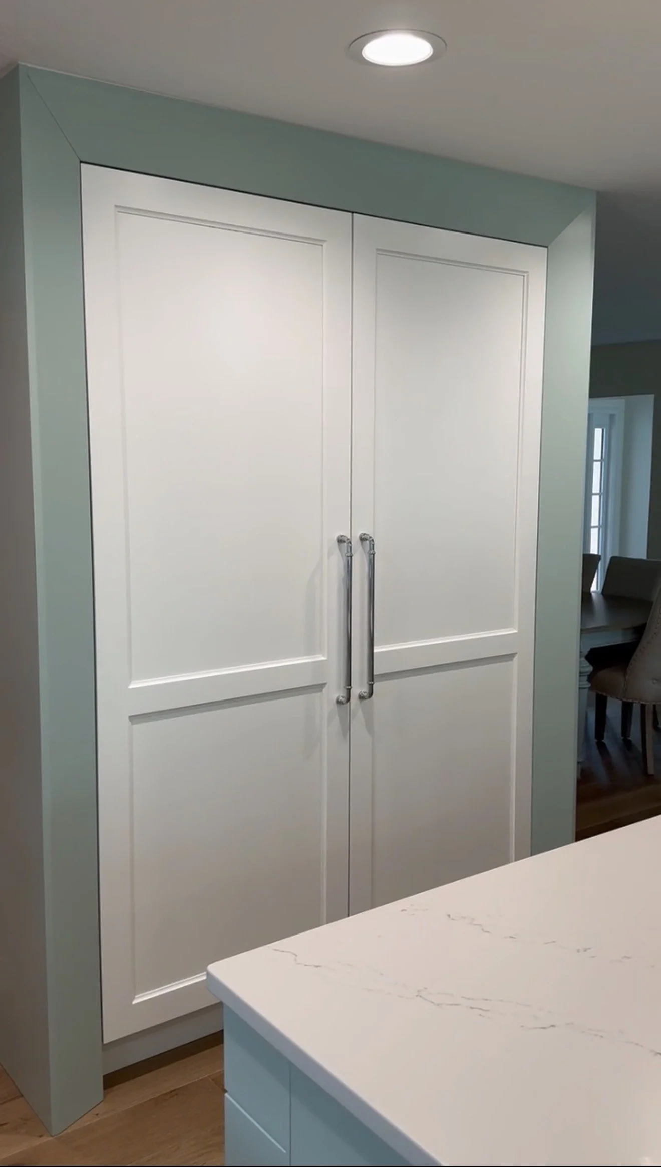 White kitchen cabinet with double doors and silver handles, marble countertop in the foreground, light green wall, ceiling light, dining area with chairs and large windows in the background.